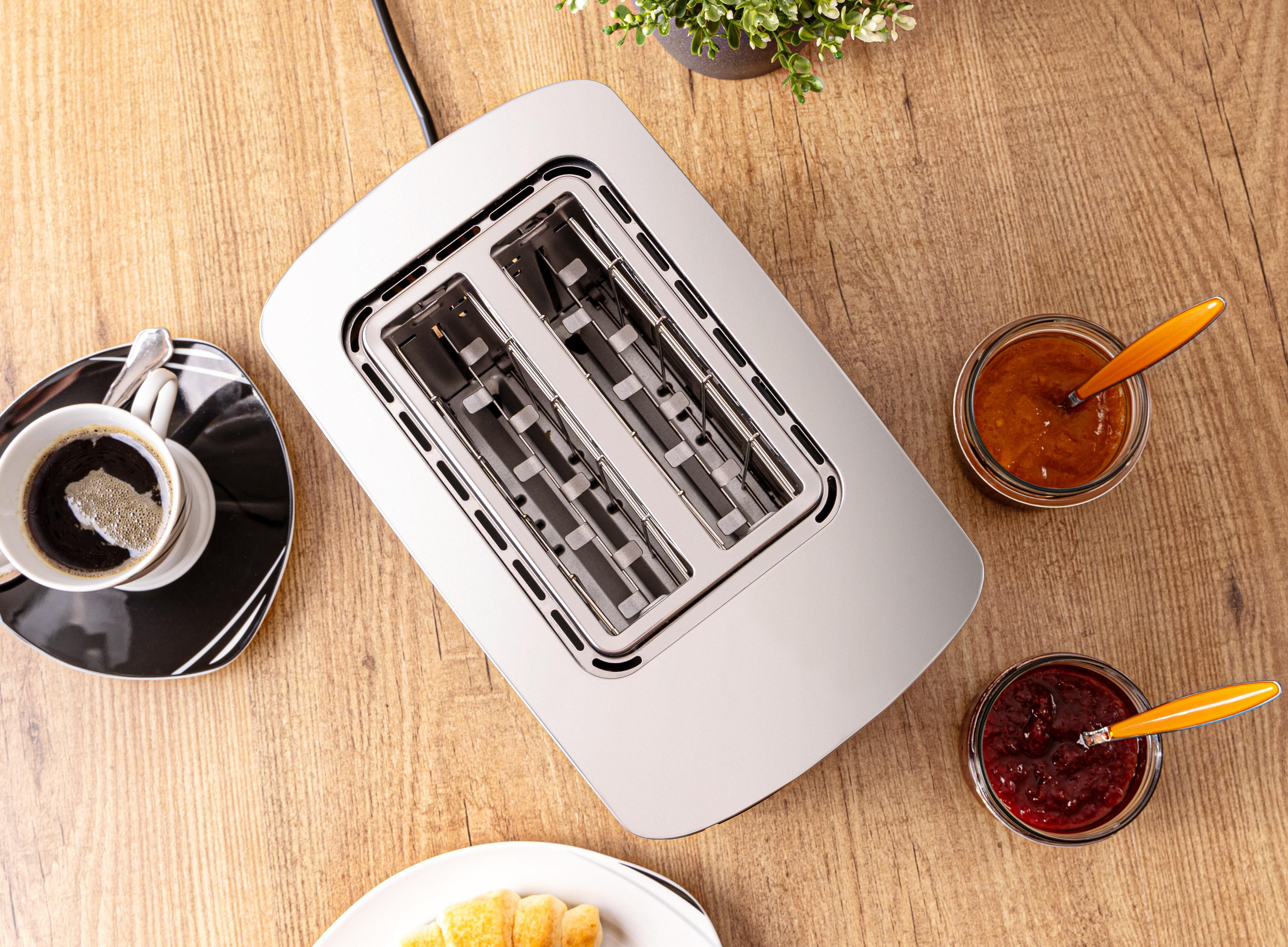 Toaster mit Brot. Zwei Gläser Marmelade. Kaffeetasse auf Untertasse. Holztisch. Draufsicht.