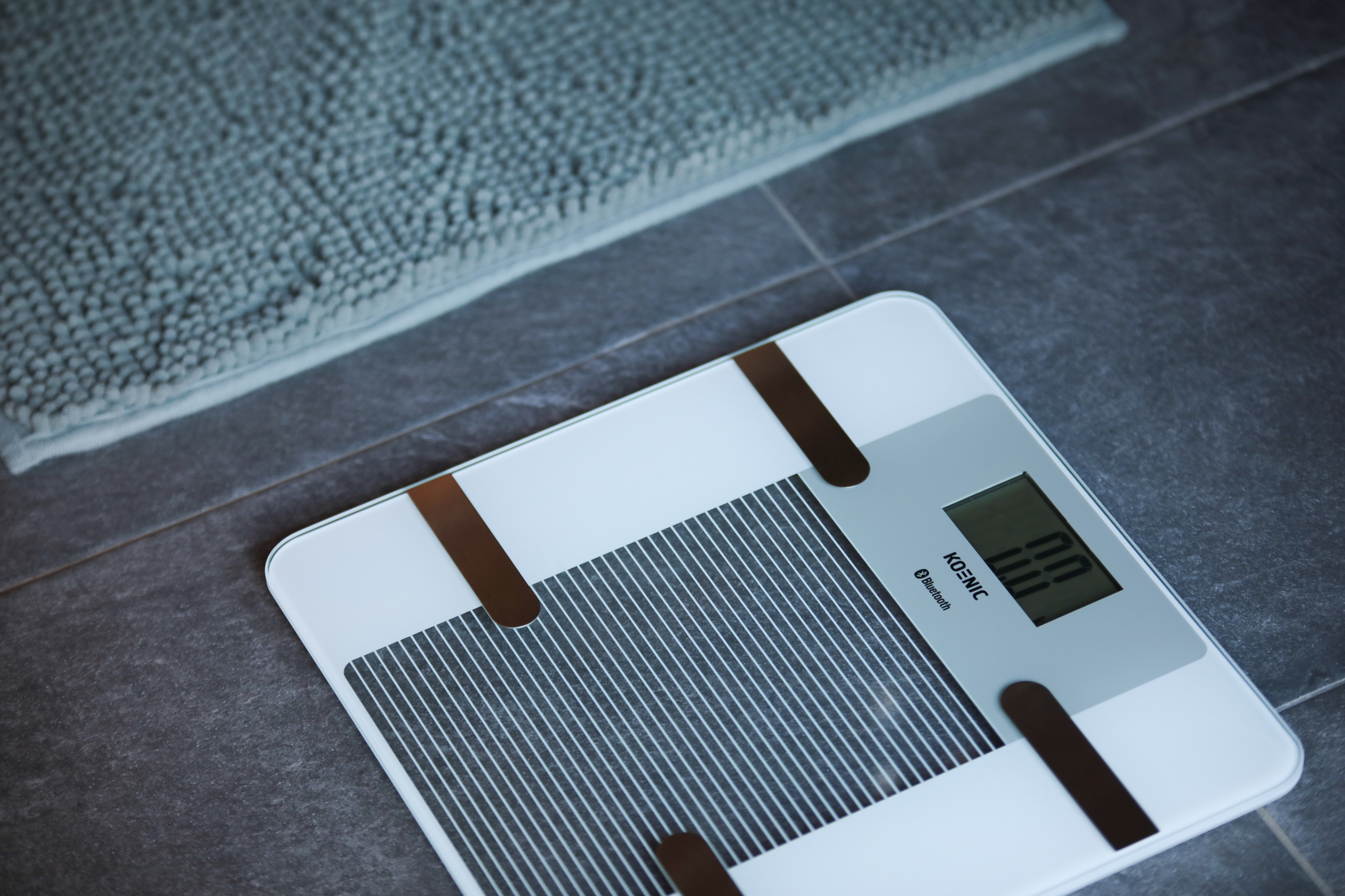 Eine Digitalwaage steht auf dem Badezimmerboden neben einer Badematte.