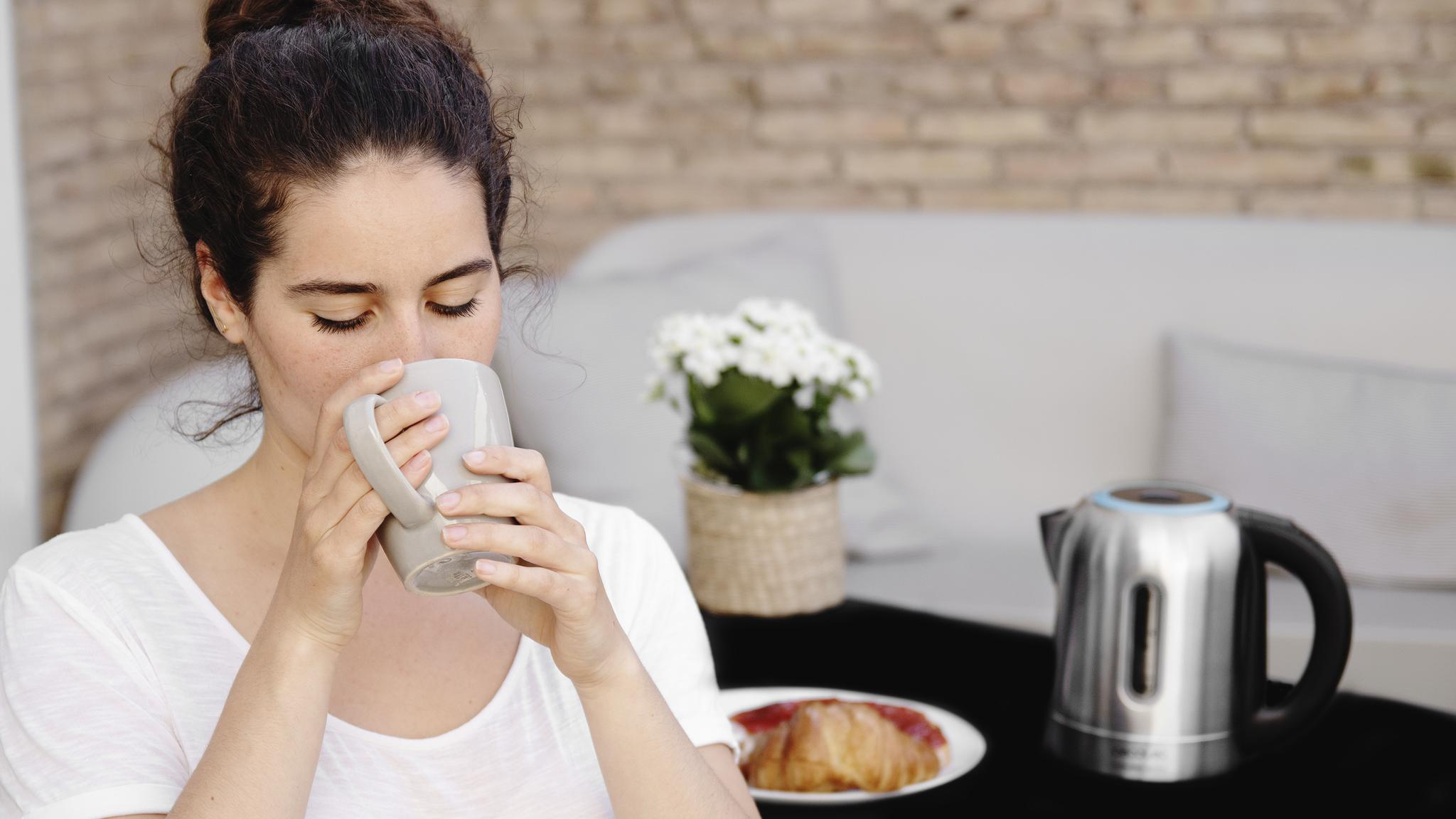 Mujer bebiendo de una taza. Mesa con comida y tetera. Fondo sofá y plantas.