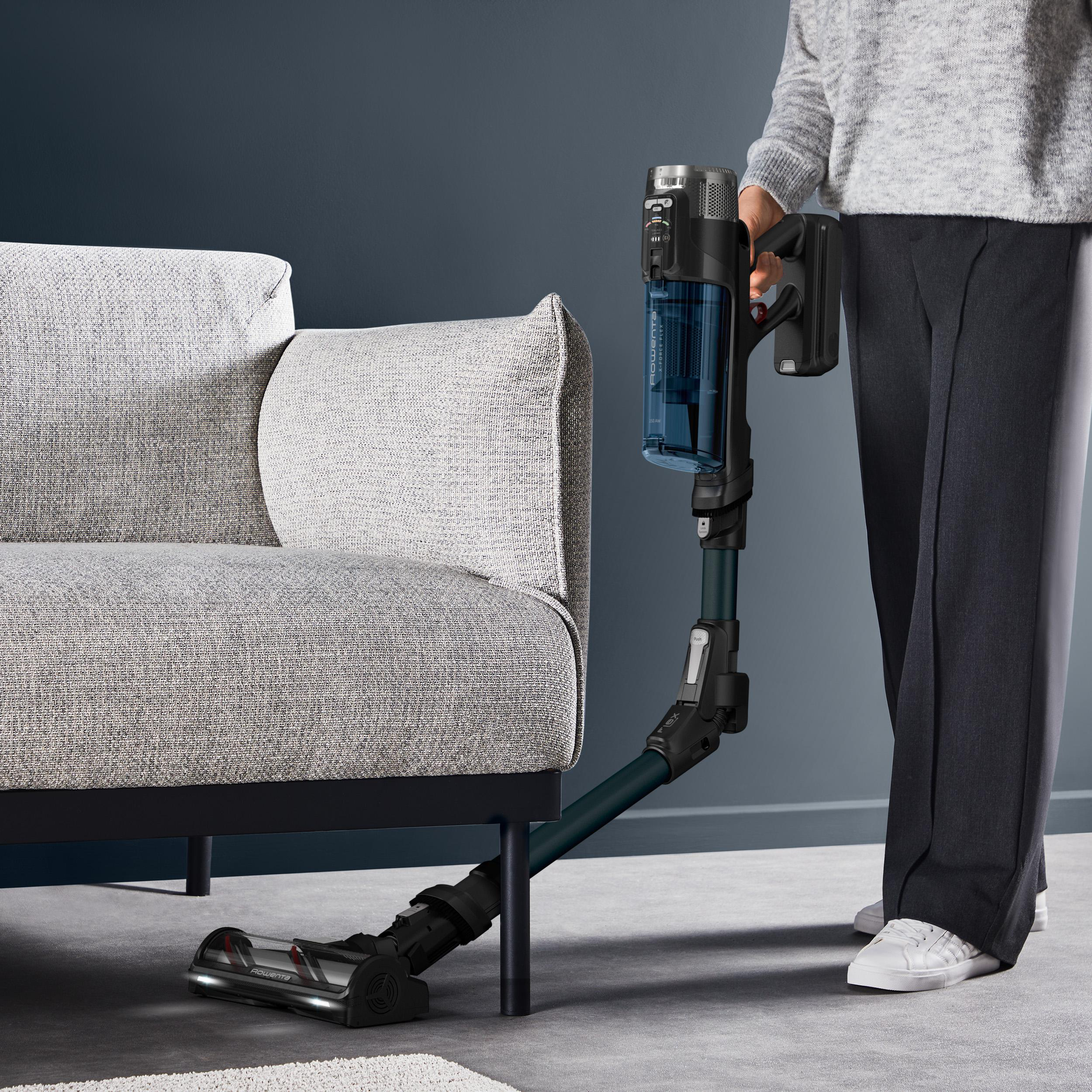 Person mit einem schwarz-blauen Staubsauger, der ein graues Sofa reinigt. Der Hintergrund ist eine dunkelblaue Wand.