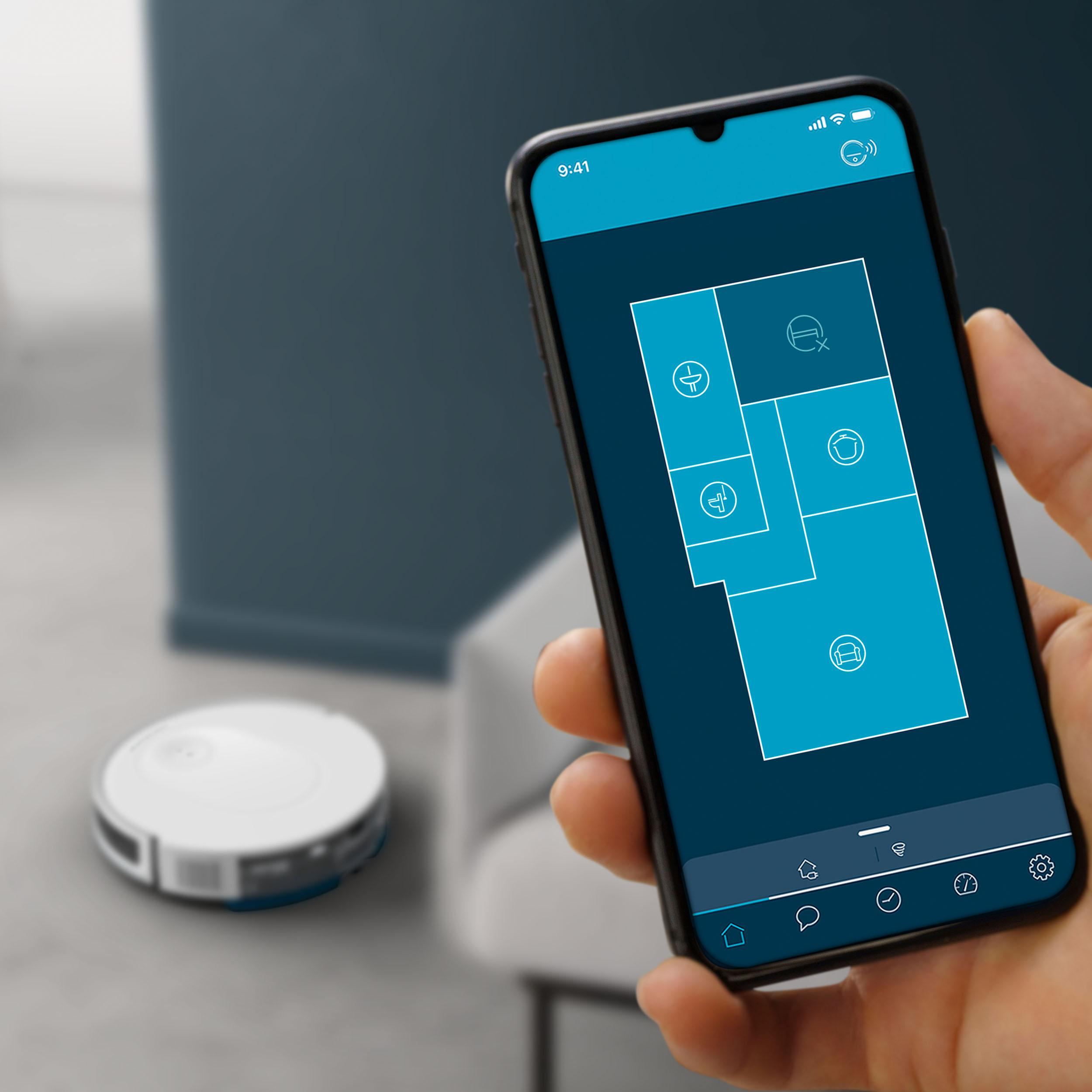 Handy mit Raumlayout-Anzeige. Ein Roboter-Staubsauger ist im unscharfen Hintergrund zu sehen.