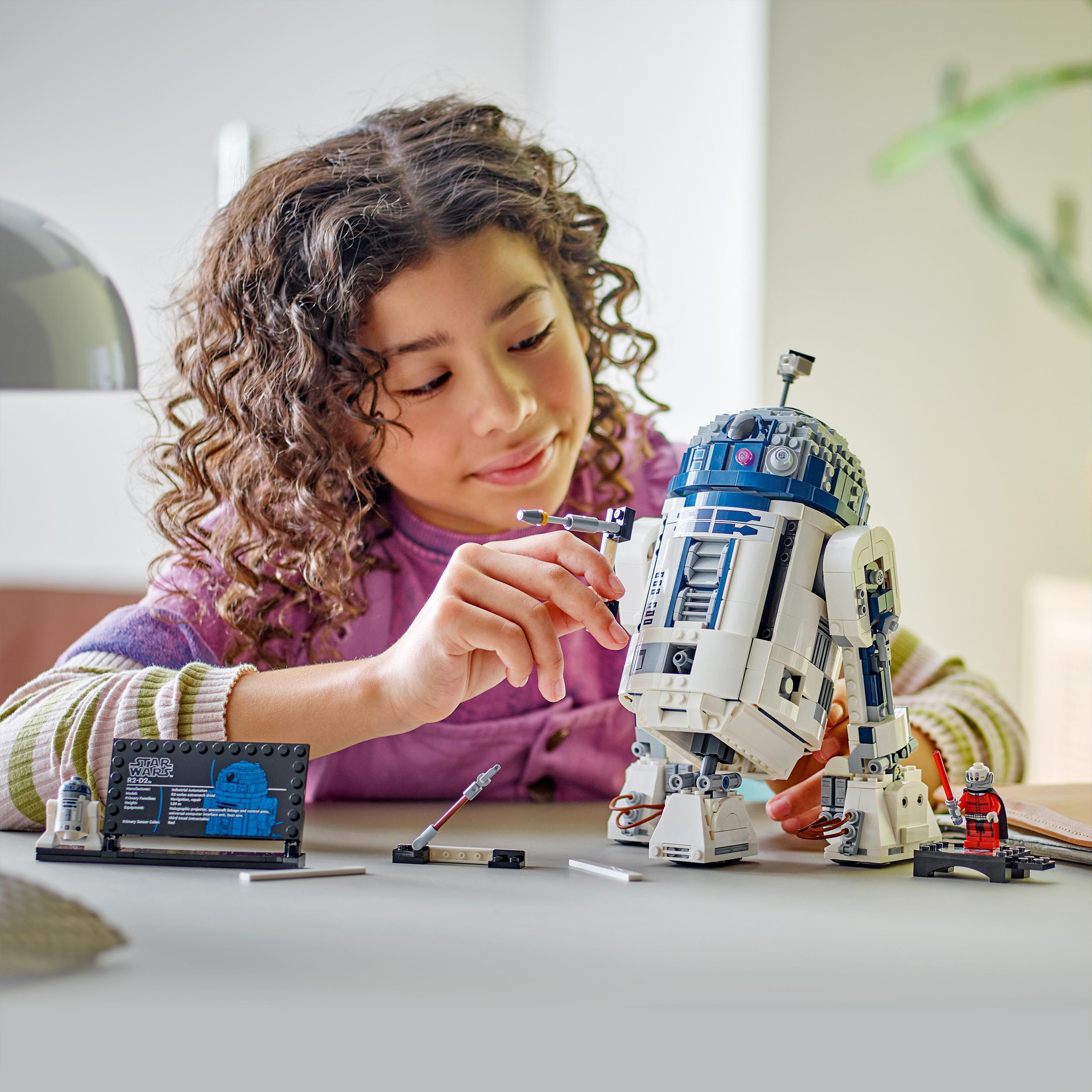 Ein Mädchen baut ein LEGO R2-D2 Modell. Der Roboter steht auf einem Tisch, Teile und Anweisungen in der Nähe.