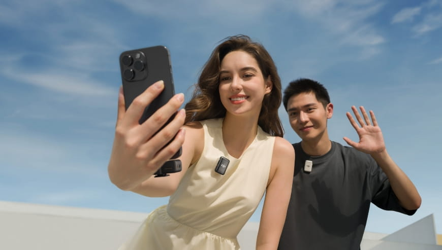 Dwie osoby robiące selfie. Jedna trzyma telefon, druga macha. Obie noszą małe mikrofony. Tło niebieskiego nieba.