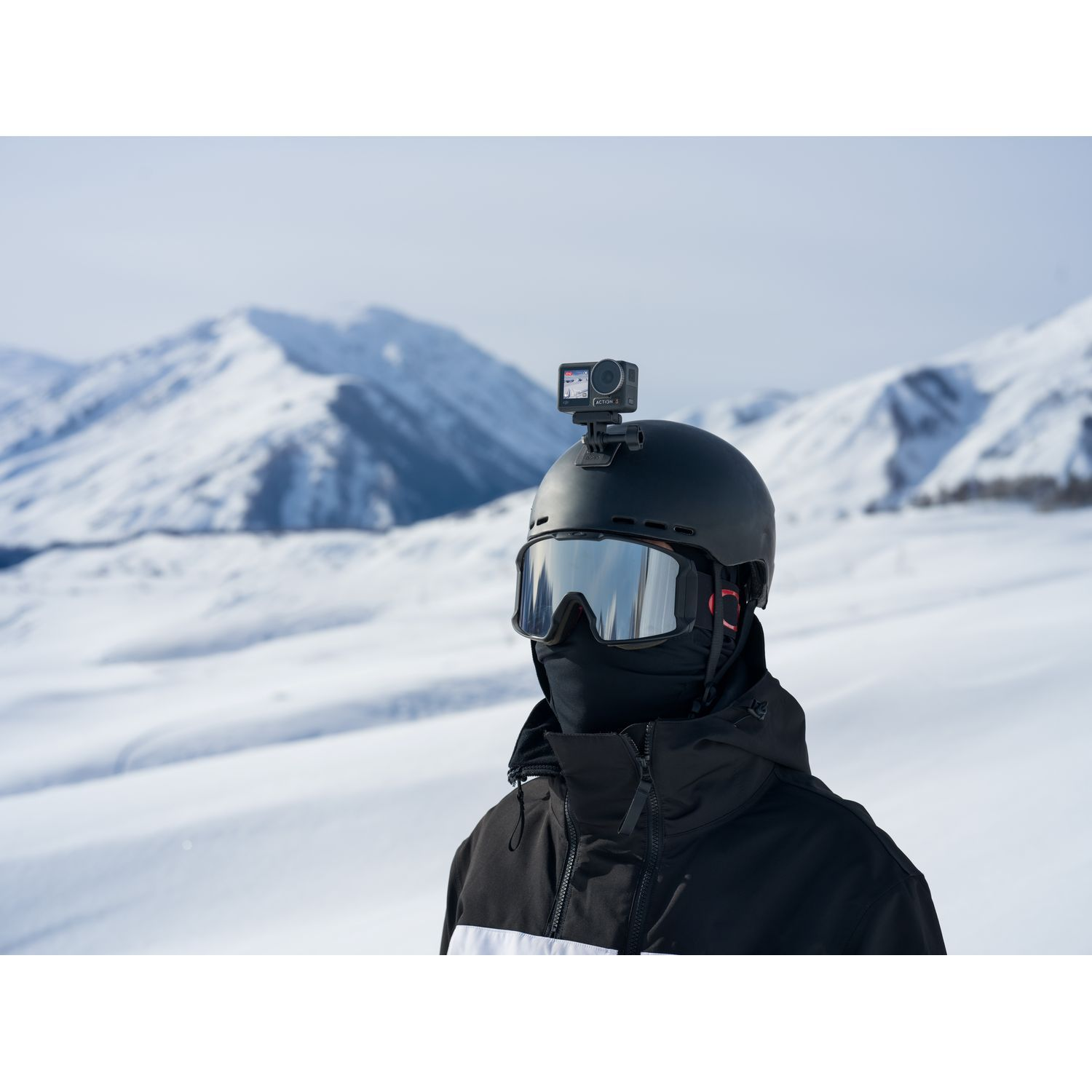 Une personne porte un casque et des lunettes noirs, avec une petite caméra montée sur le casque, de la neige en arrière-plan.