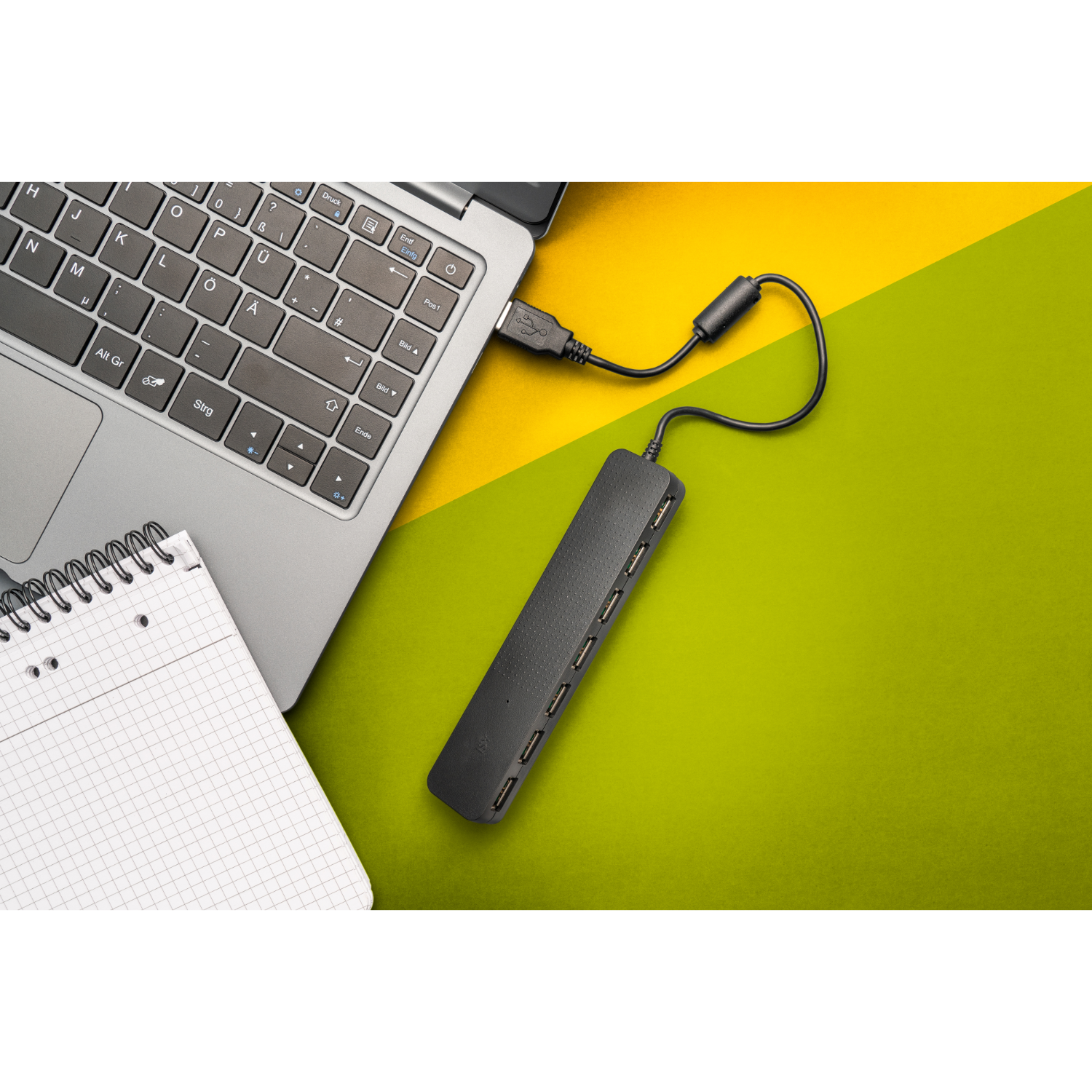 Een laptop aangesloten op een USB-hub met meerdere USB-poorten, geplaatst op een groen en geel oppervlak.