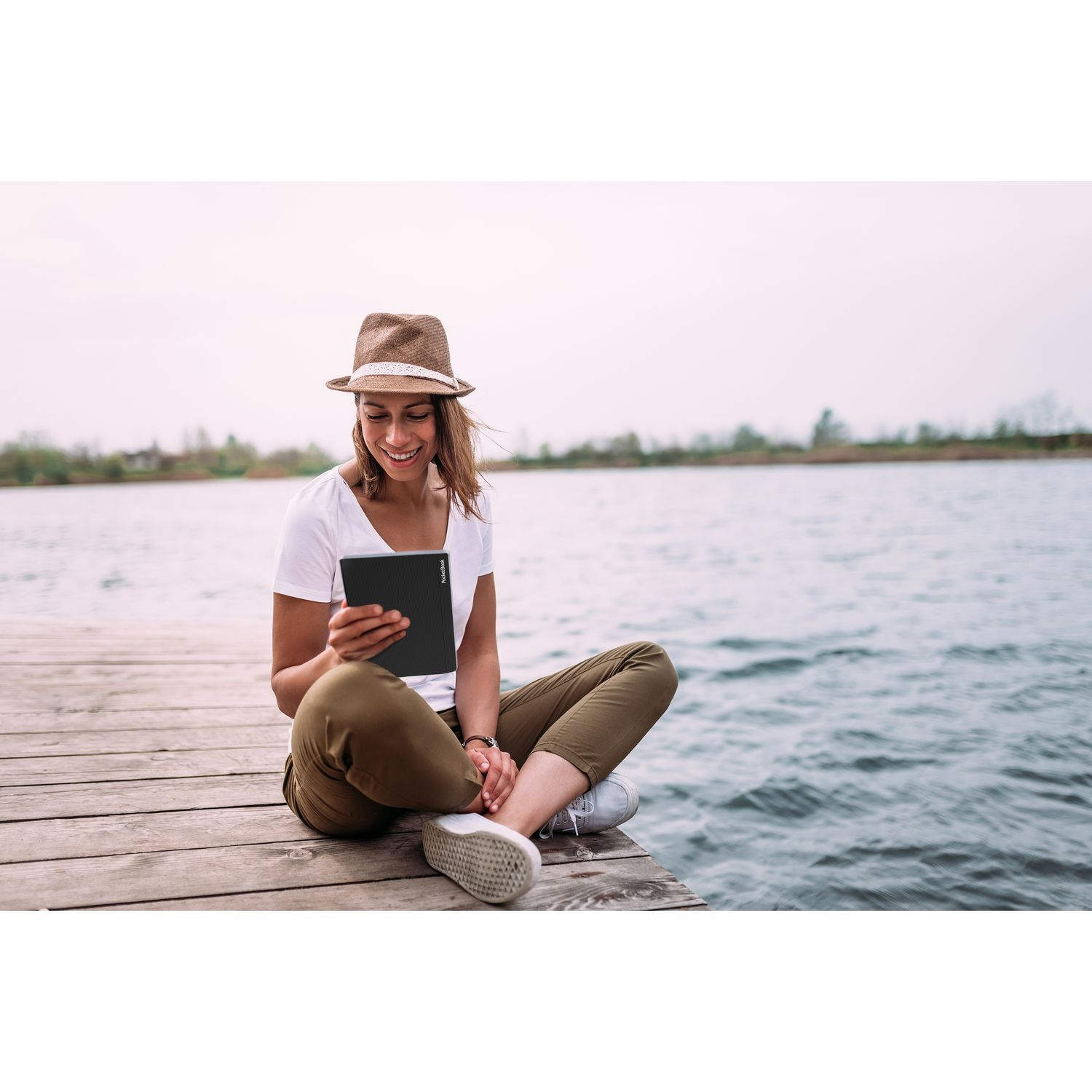 Femme assise sur un ponton, lisant une tablette. Lac en arrière-plan, ciel nuageux.