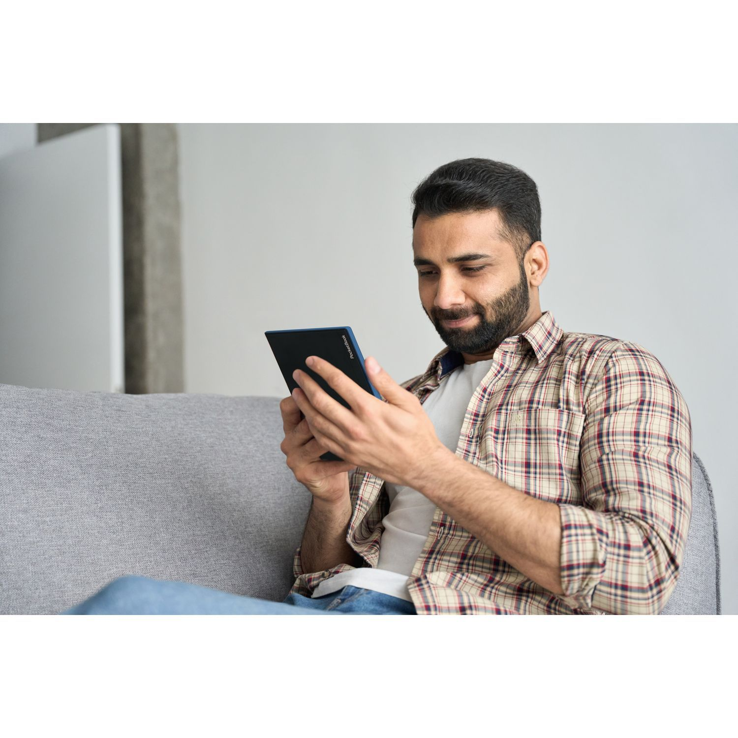 Homme assis sur un canapé, regardant une tablette noire à deux mains. Il sourit.
