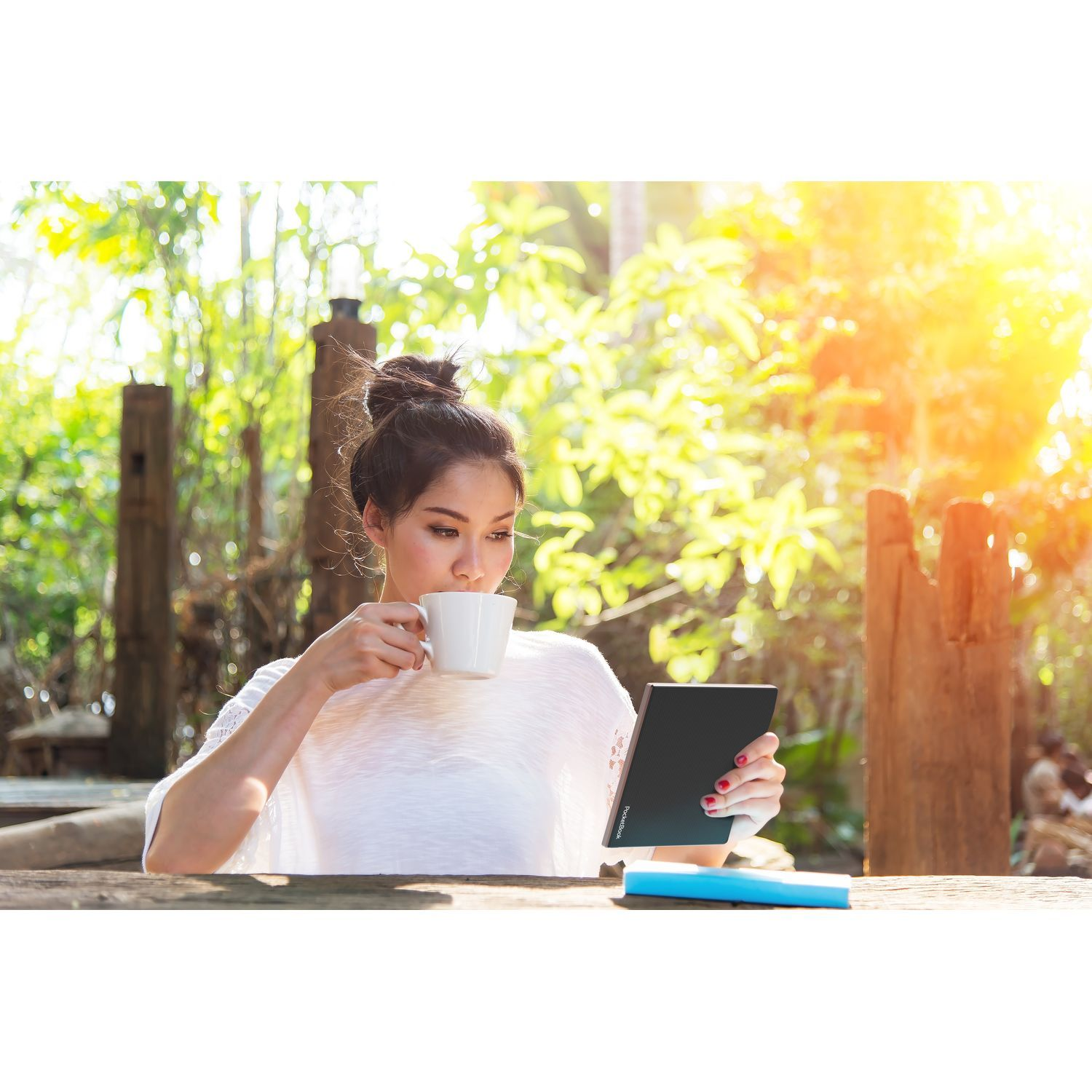 Une femme boit dans une tasse tout en tenant une tablette. Elle est assise dehors dans un environnement ensoleillé.