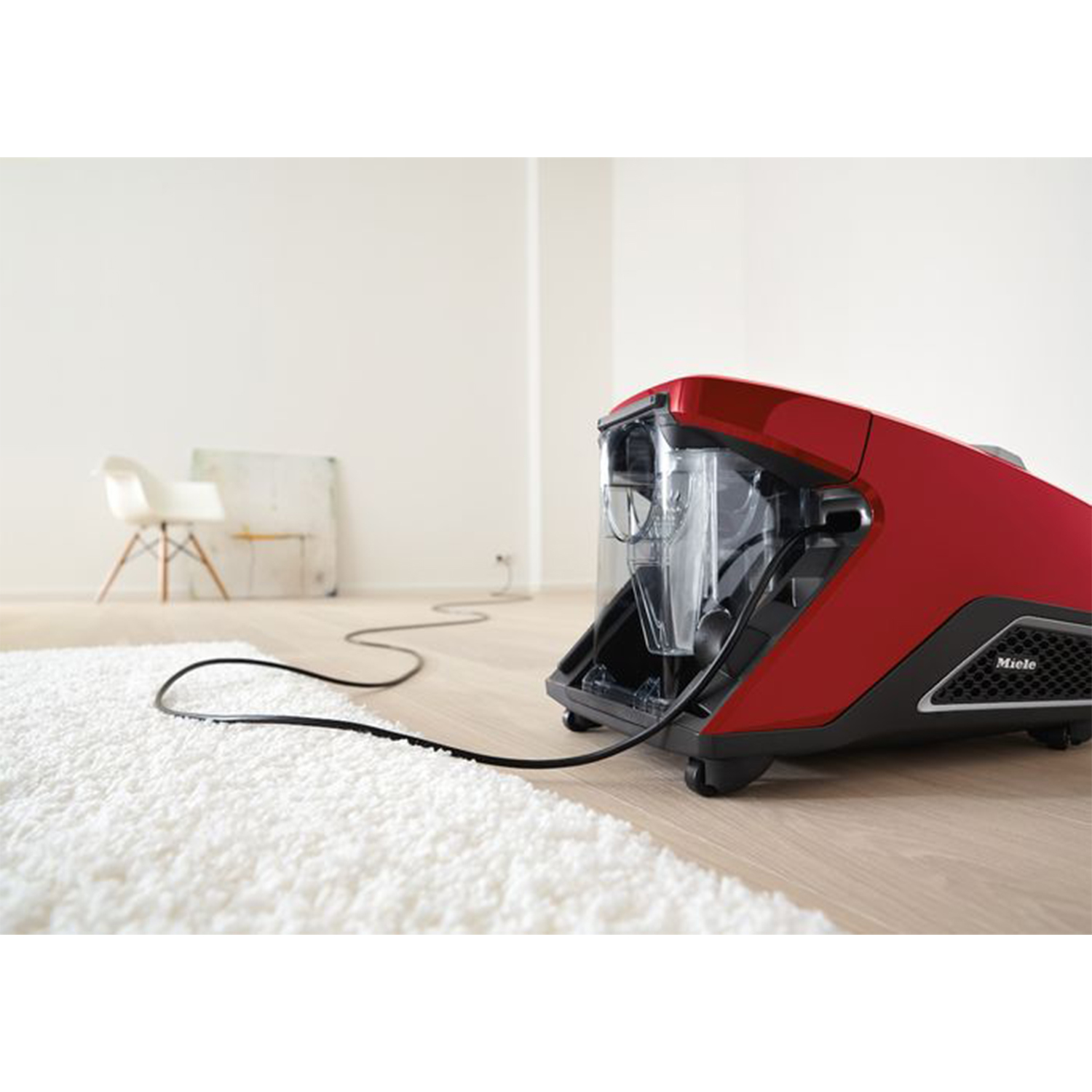 Aspirapolvere rosso con contenitore trasparente su pavimento in legno con tappeto bianco.