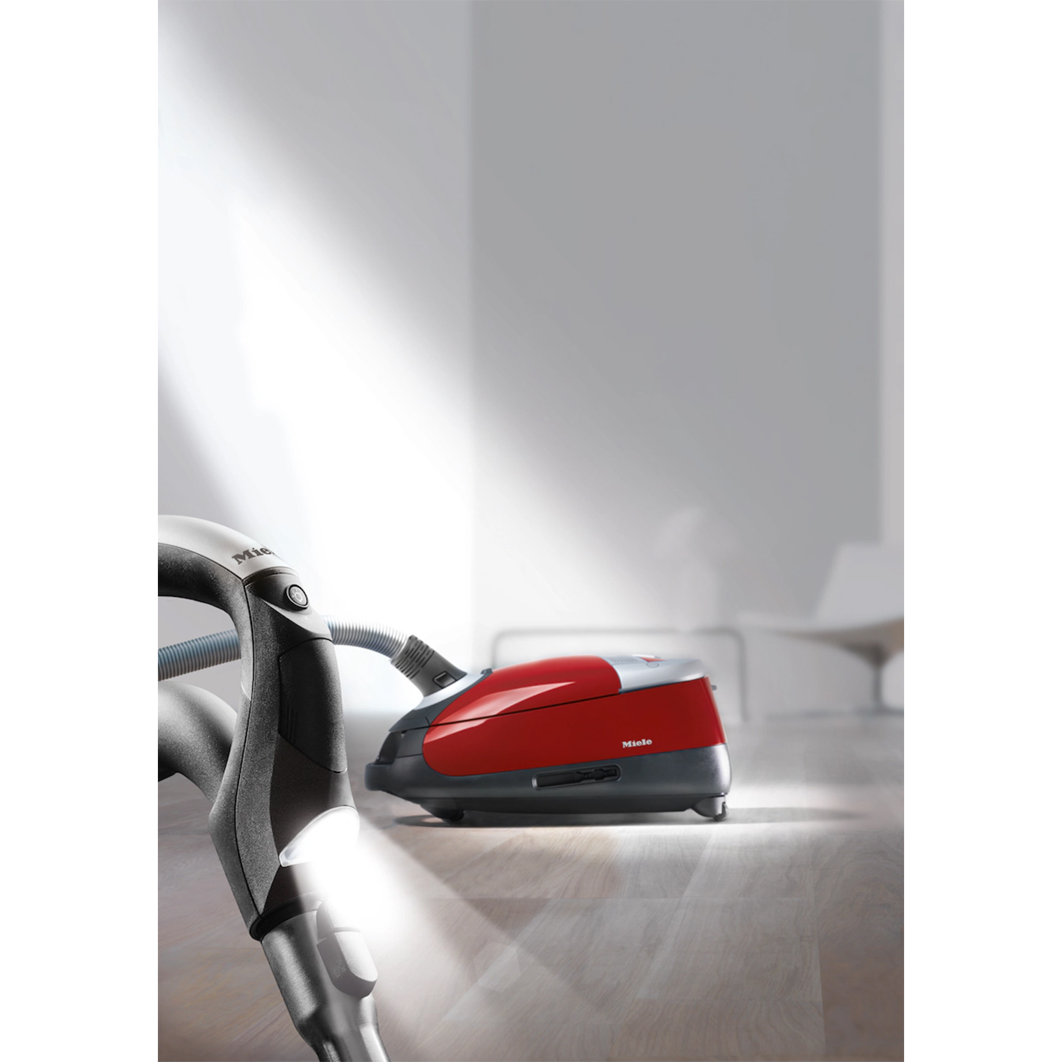 Aspirapolvere Miele rosso e grigio su pavimento in legno chiaro, con un fascio di luce.