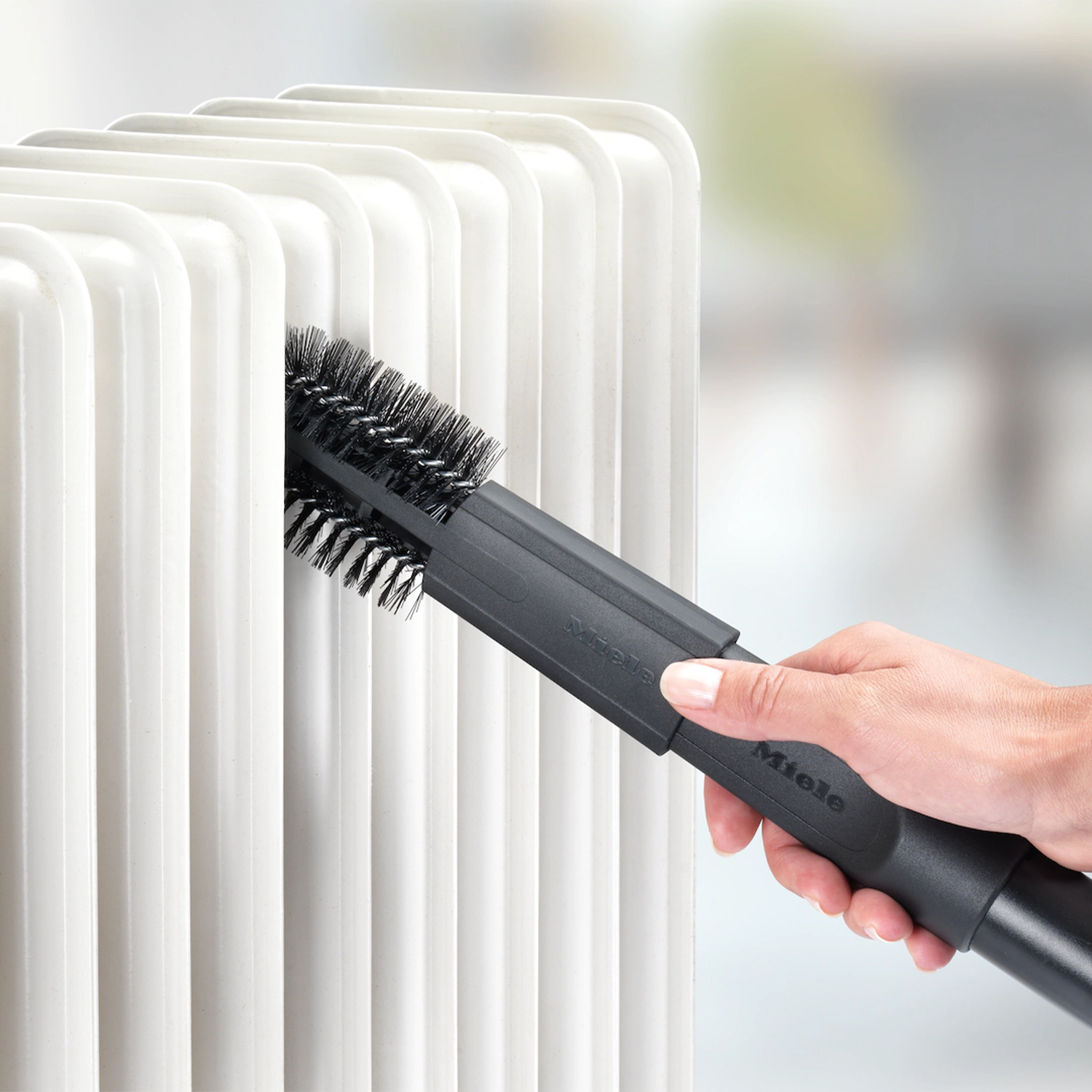 Una mano che pulisce un radiatore bianco con una spazzola nera attaccata a un aspirapolvere.