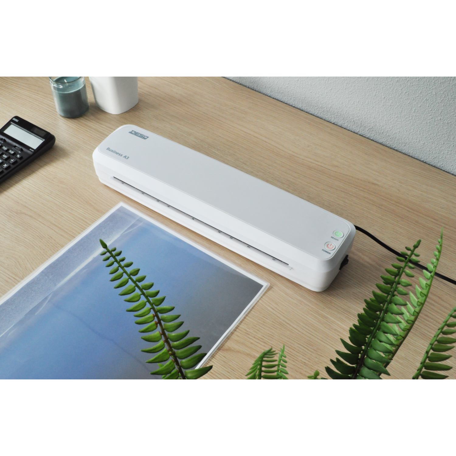 Laminator machine on a wooden table with a fern and a calculator.