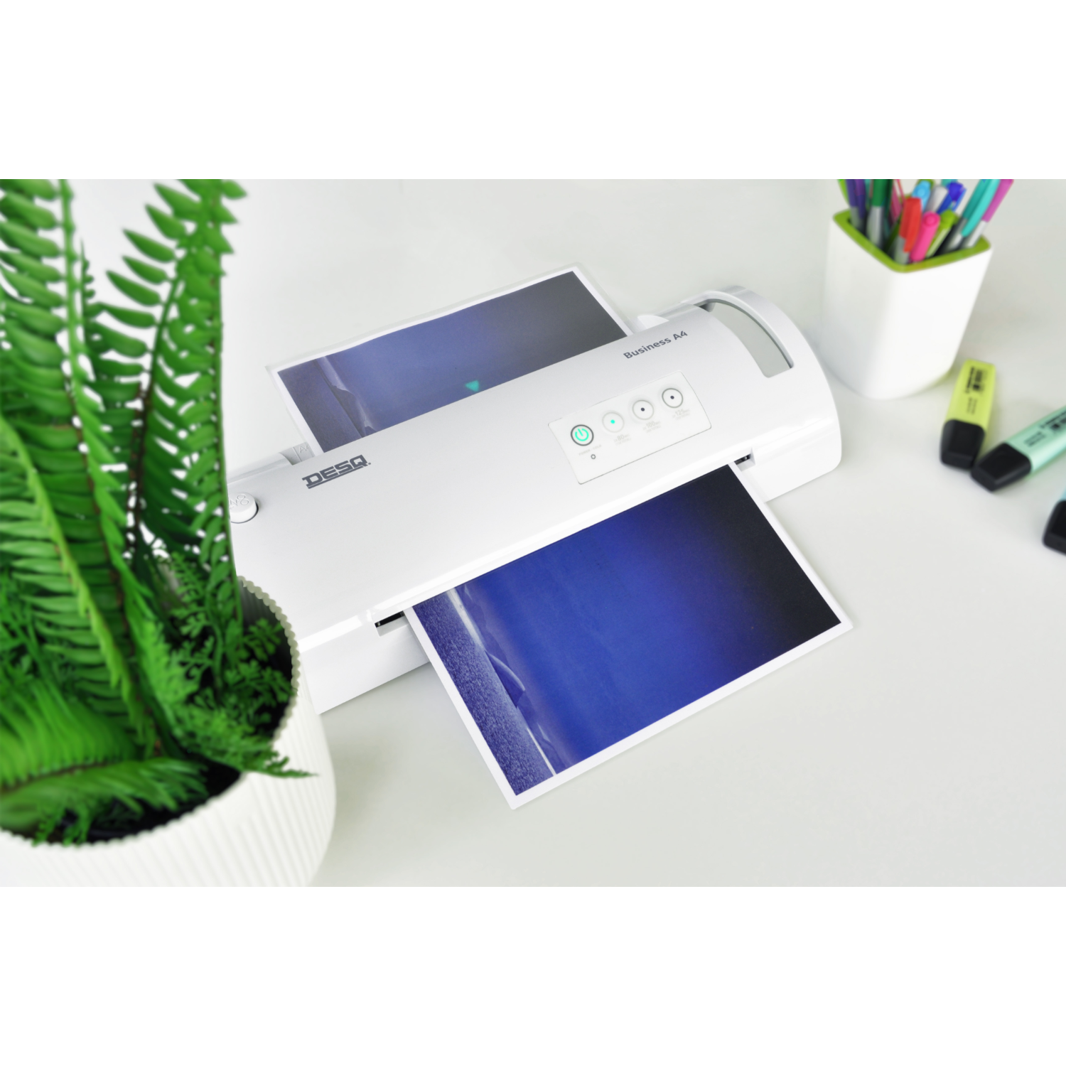 A laminator with paper going through and a green plant.