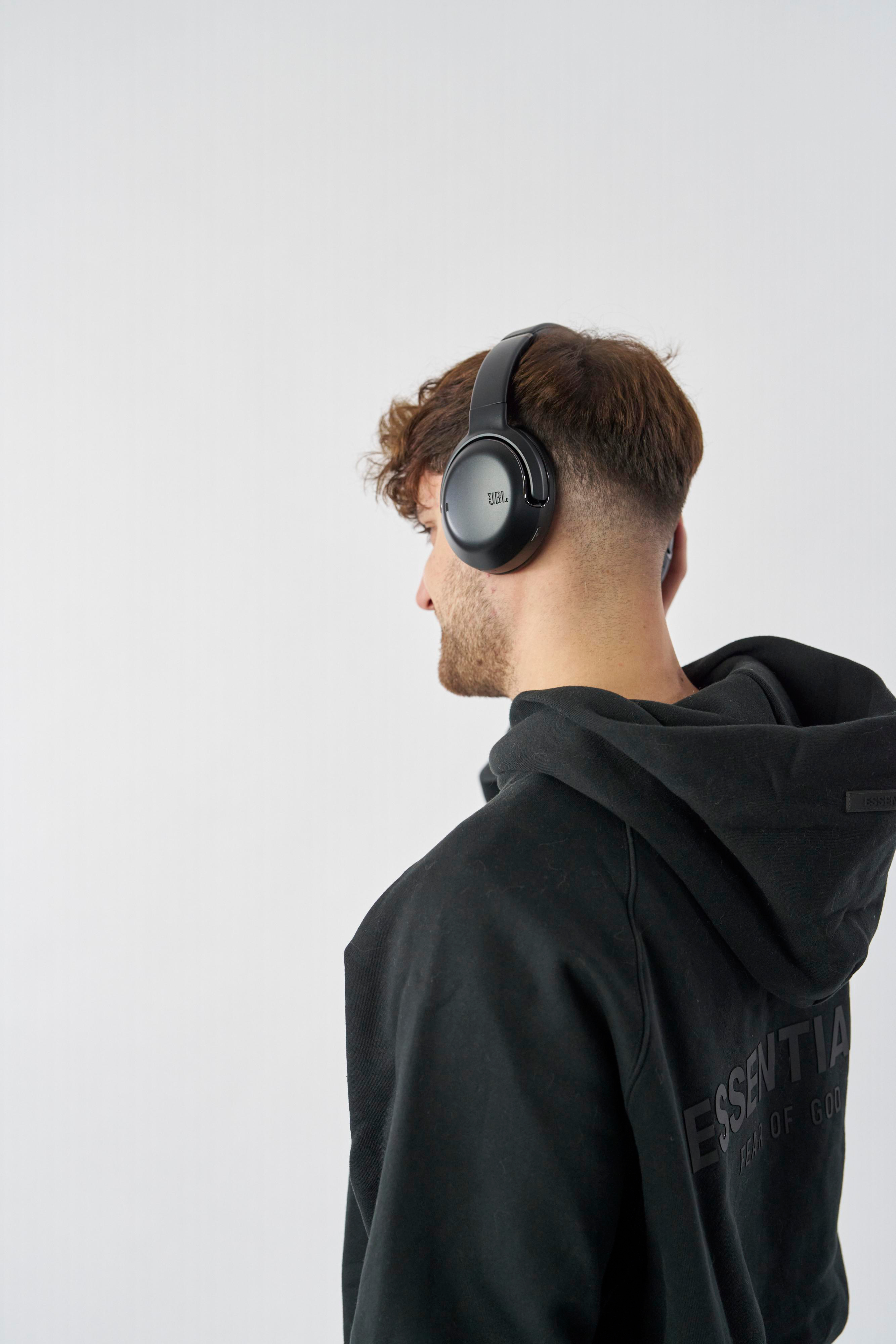 Ein Mann mit schwarzen JBL-Kopfhörern. Er trägt einen schwarzen Kapuzenpullover und schaut weg.