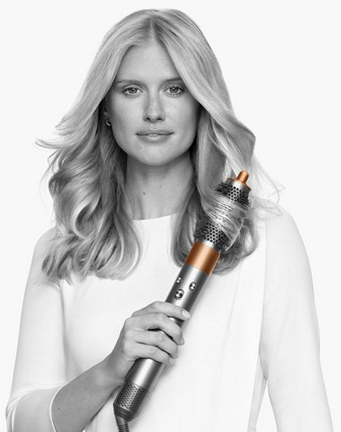 Donna bionda che tiene un'arricciacapelli Dyson argento e rame, con lunghi capelli mossi.