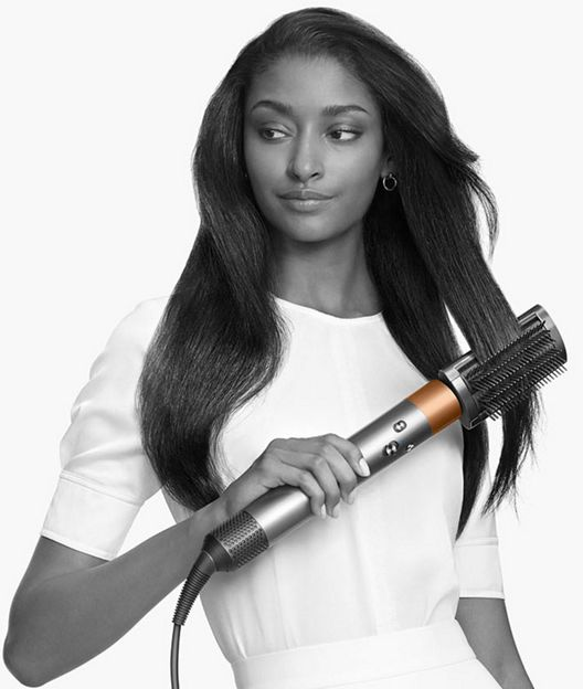 Una donna con lunghi capelli scuri tiene in mano uno strumento per lo styling dei capelli Dyson color argento e rame.