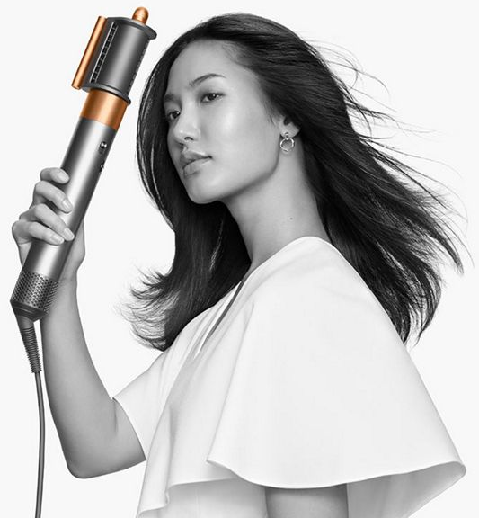 Una donna tiene un arricciacapelli argento e rame. Ha i capelli scuri e indossa un top bianco.