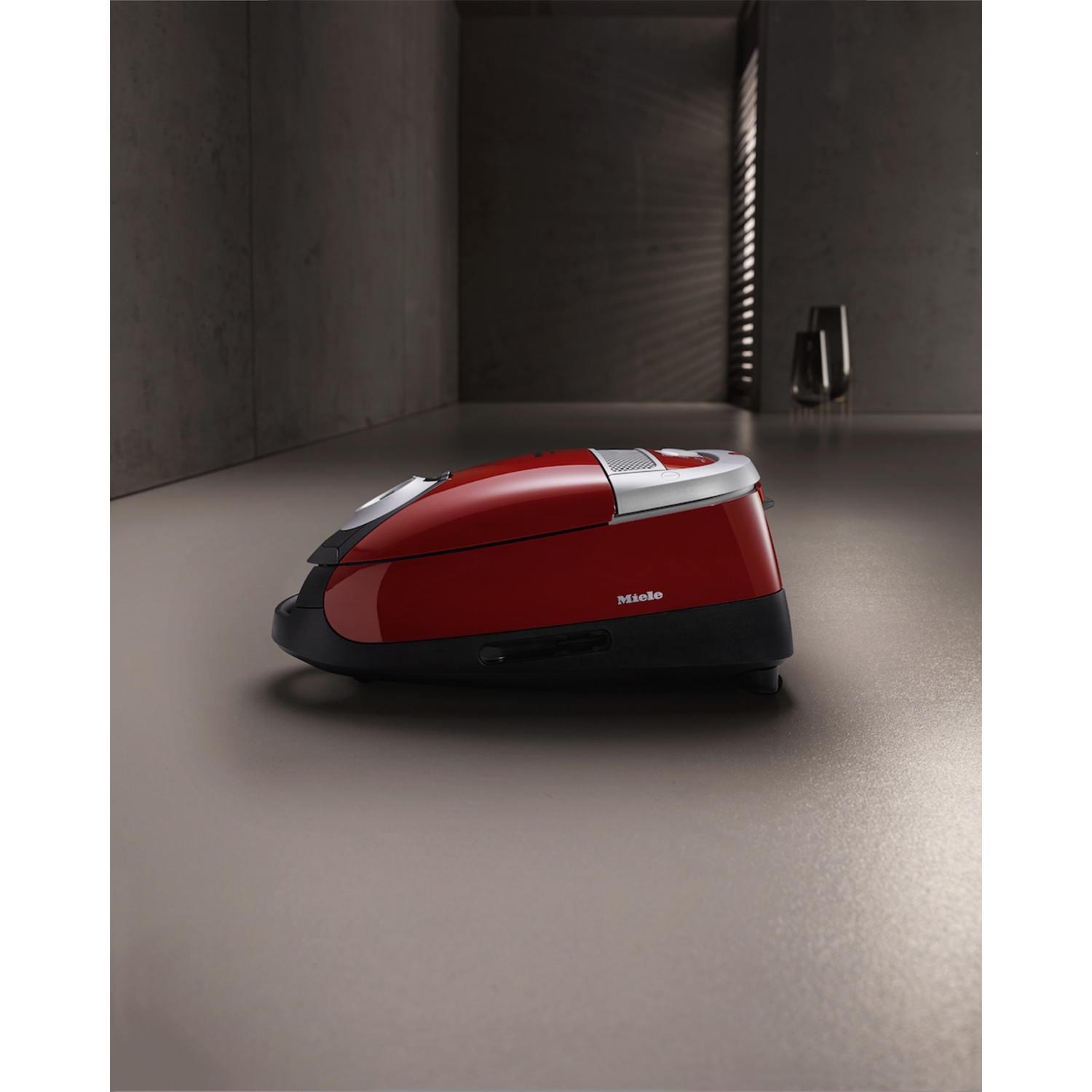 Un aspirapolvere rosso su una superficie grigia con sfondo scuro, etichettato Miele.