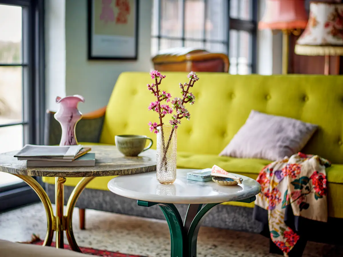 Salon z żółtą sofą, dwoma małymi stolikami, wazonem z kwiatami i oprawioną sztuką na ścianie.