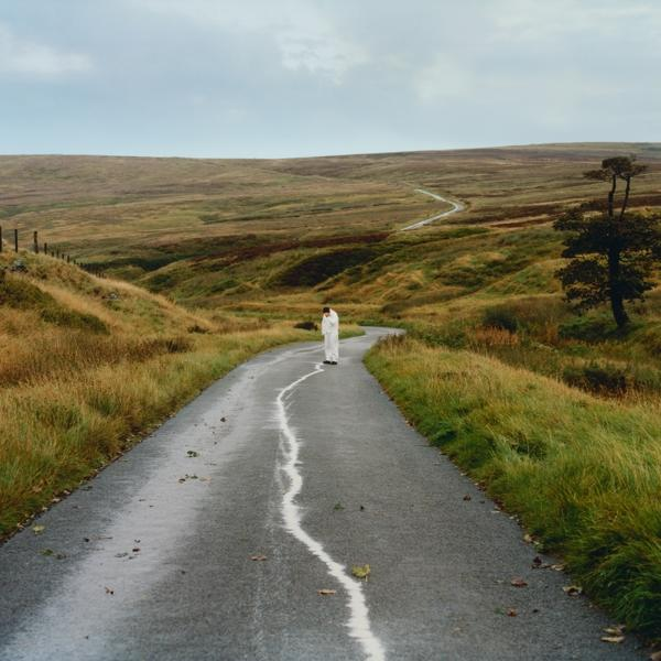 Eine Person in weißer Kleidung steht auf einer nassen Straße in einer ländlichen Landschaft.
