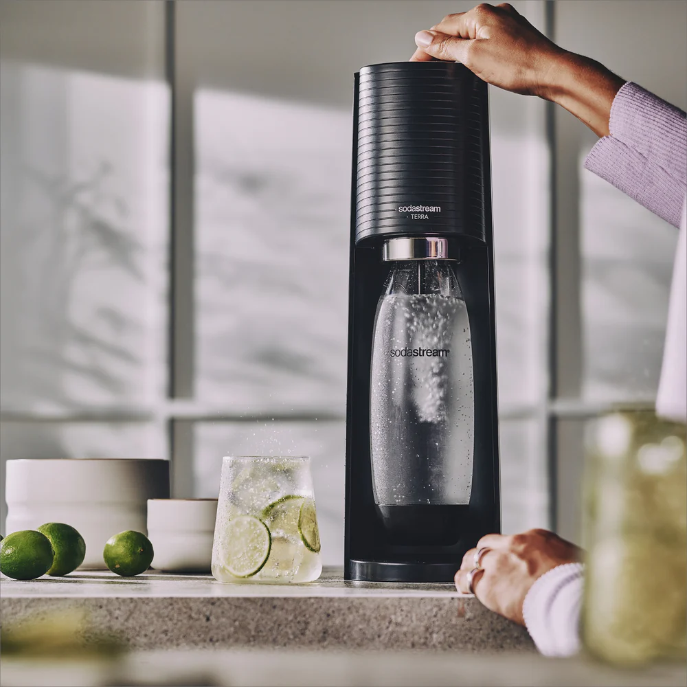 Schwarzer SodaStream mit Flasche, Glas Wasser mit Limettenscheiben und Limetten auf der Theke.