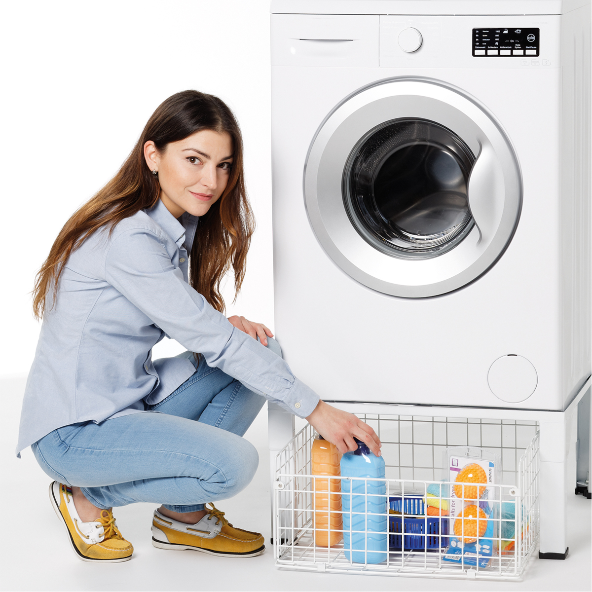 Kobieta obok białej pralki. Kuca, sięgając po produkty do prania w drucianym koszu.