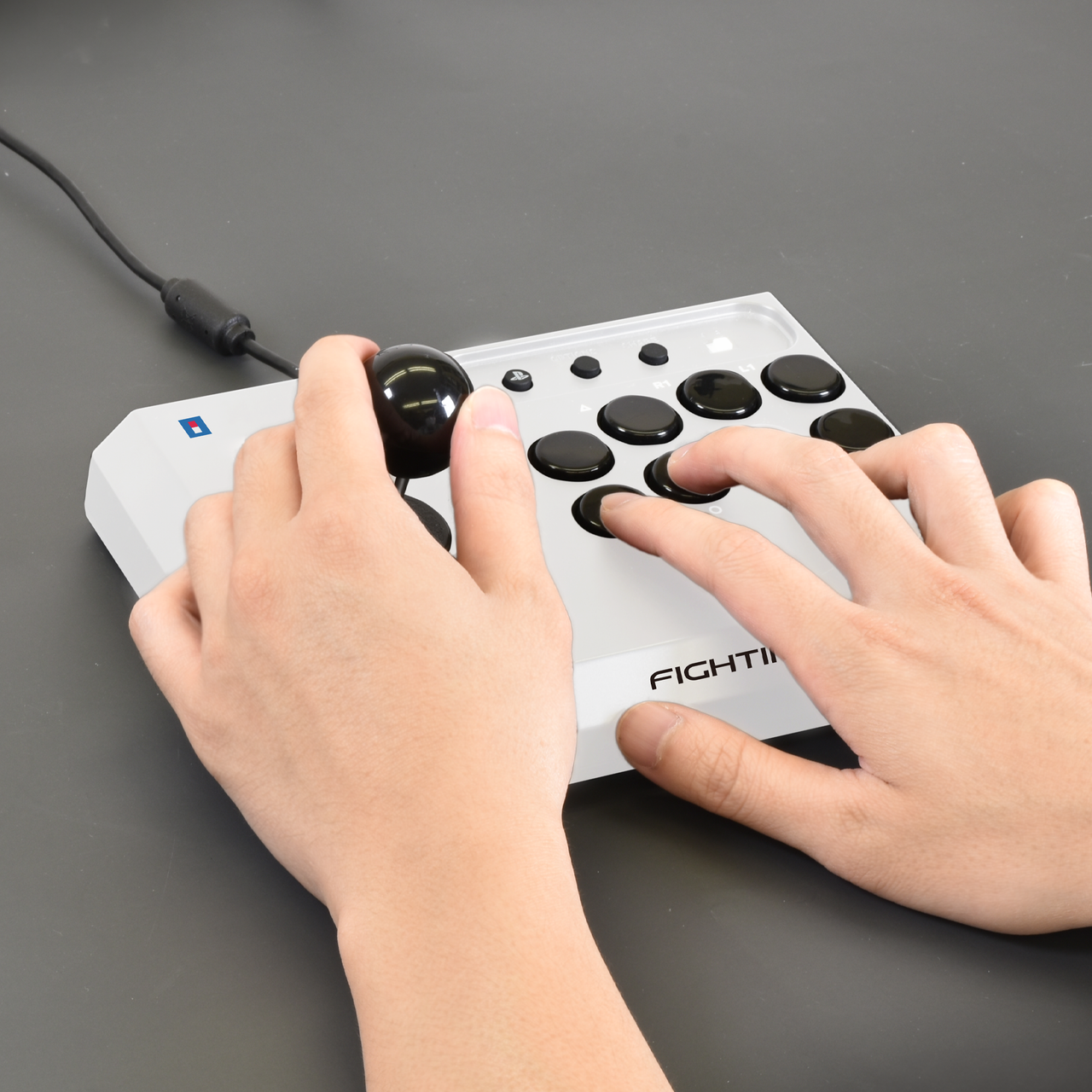 Mains utilisant une manette FIGHT blanche avec boutons noirs et joystick sur surface grise.