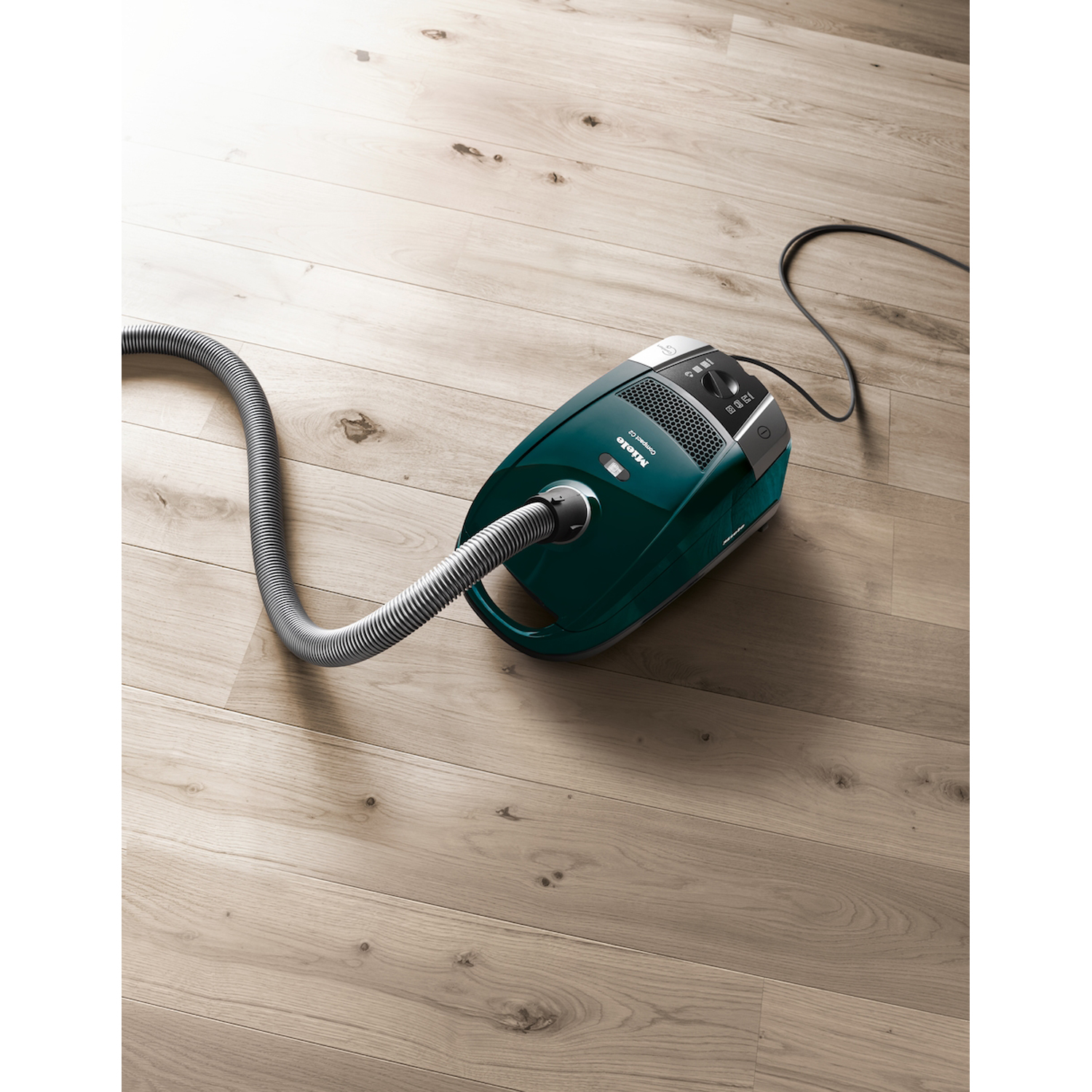 Un aspirapolvere verde con tubo grigio su pavimento in legno marrone chiaro.