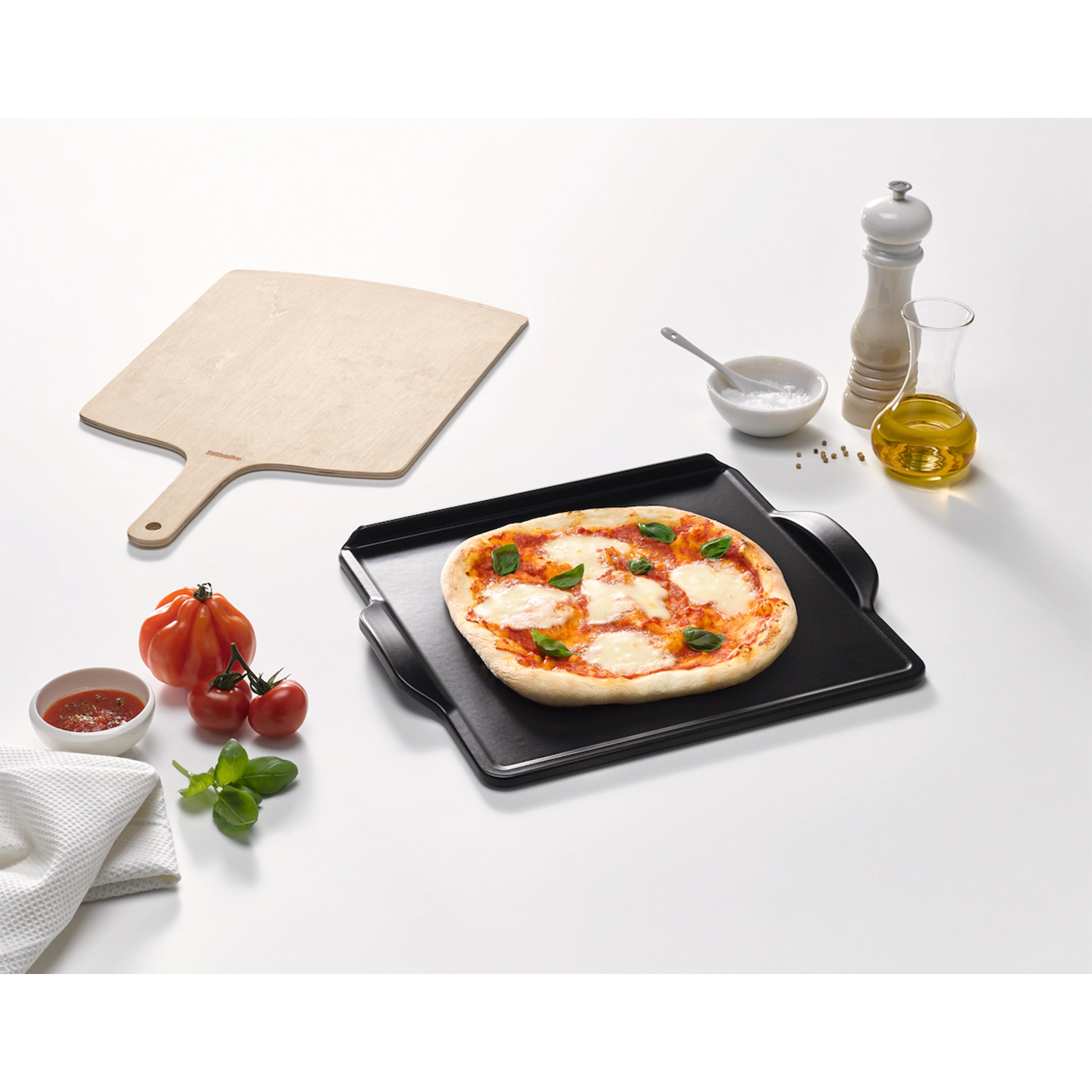 Pizza su un vassoio nero con basilico e pomodori. Pala per pizza in legno e ingredienti su tavolo bianco.