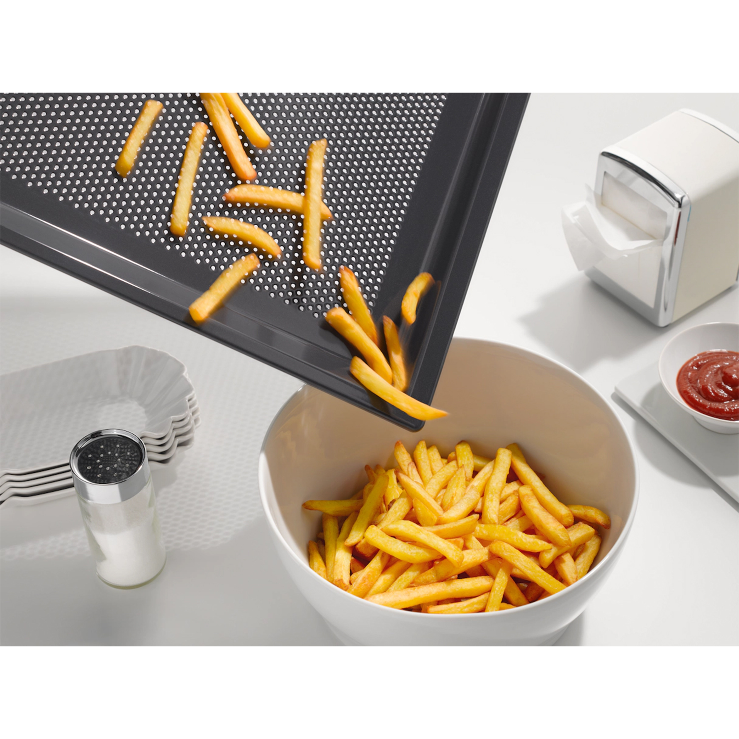 Patatine fritte versate da un vassoio nero in una ciotola bianca. Saliera e ketchup accanto.