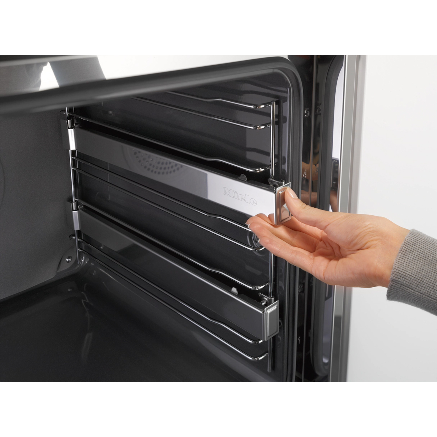 Mano che apre la griglia del forno in un forno in acciaio inossidabile. Logo Miele visibile. Interno scuro.
