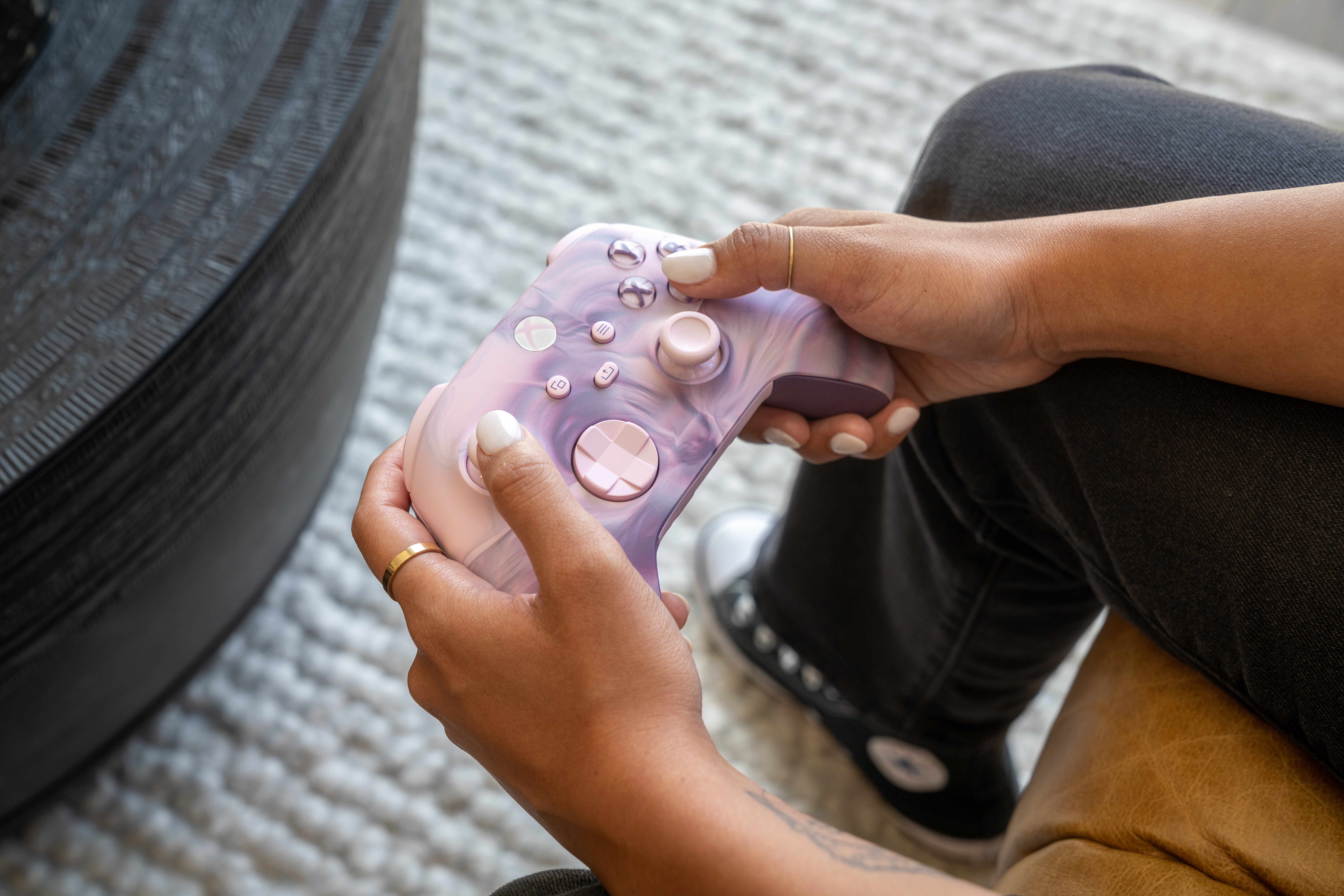 Eine Person hält einen rosa-lila Xbox-Controller. Sie trägt Jeans und Ringe. Ein schwarzer Tisch und ein weißer Teppich sind im Hintergrund.