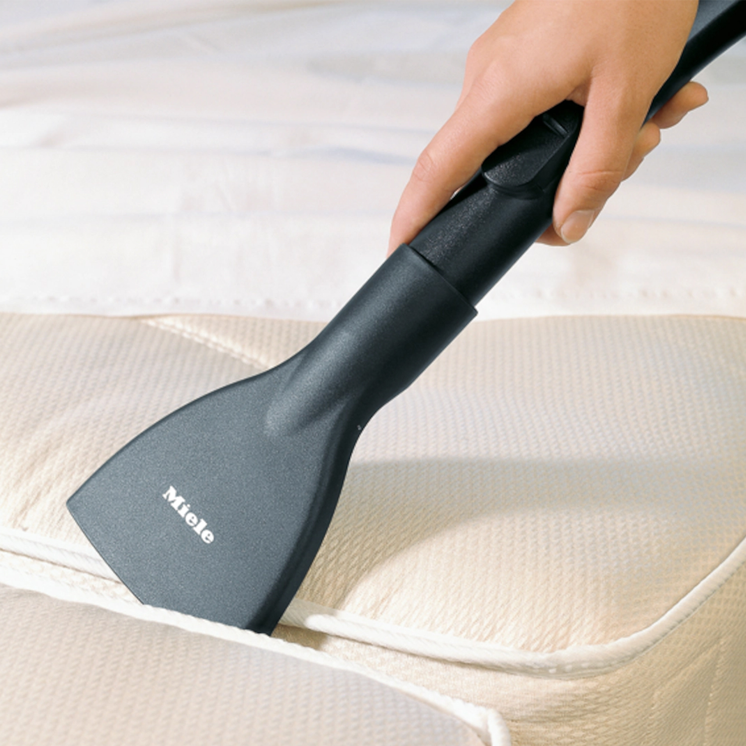 Una mano aspira un materasso con un aspirapolvere Miele. Il materasso è beige.