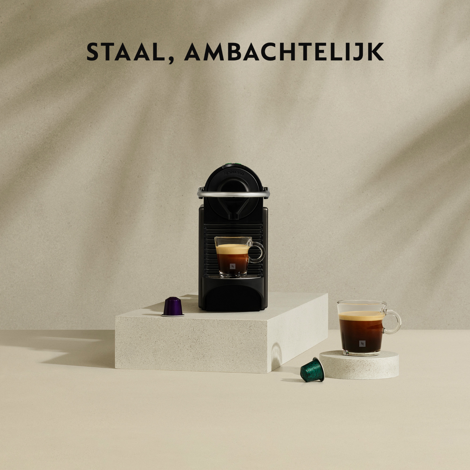 Zwarte Nespresso machine met een kopje koffie, twee capsules en een glas koffie op een oppervlak.