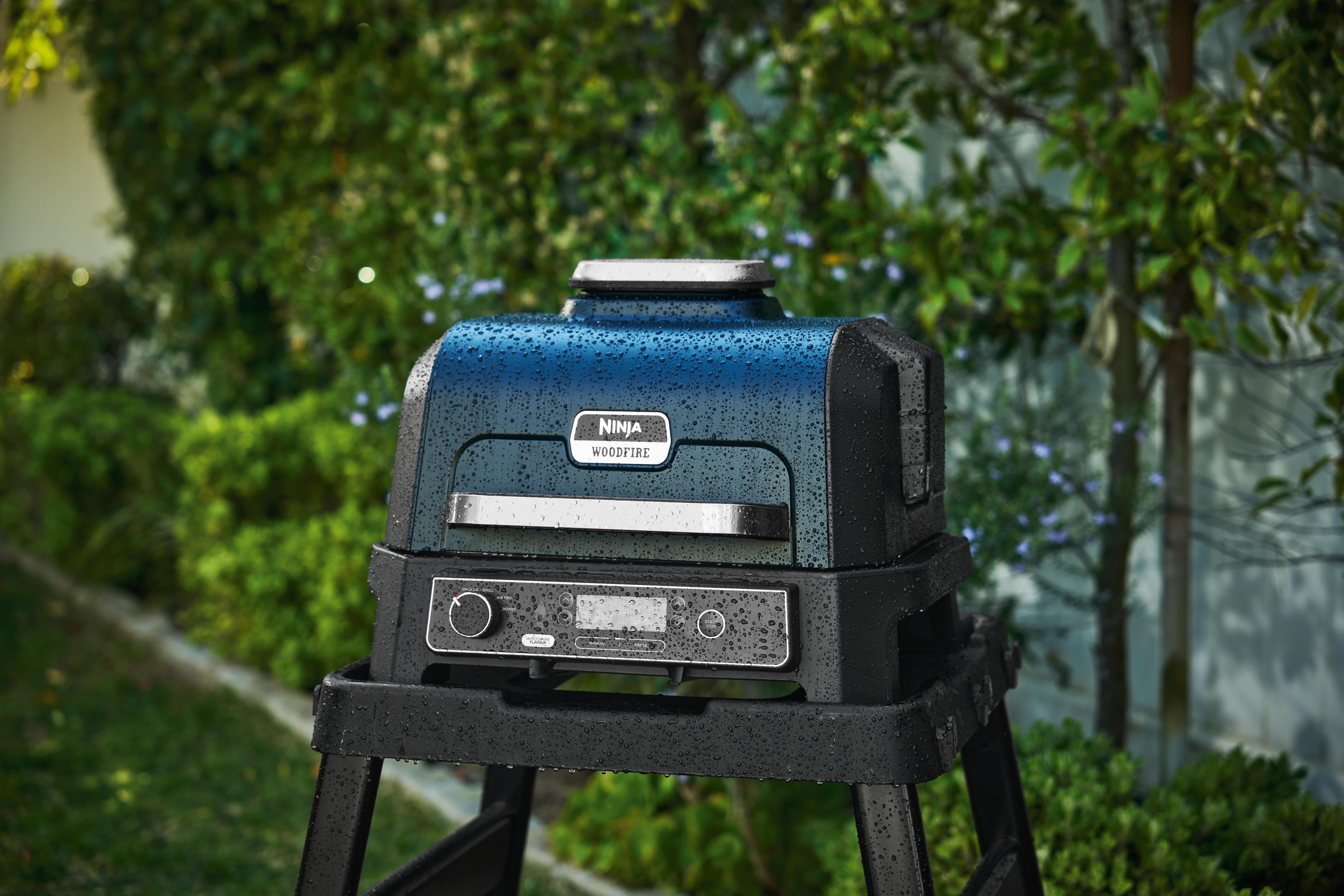 Ein schwarzer und blauer Ninja Woodfire Grill auf einem Ständer, in einem Garten mit grünen Büschen und einem Baum im Hintergrund.