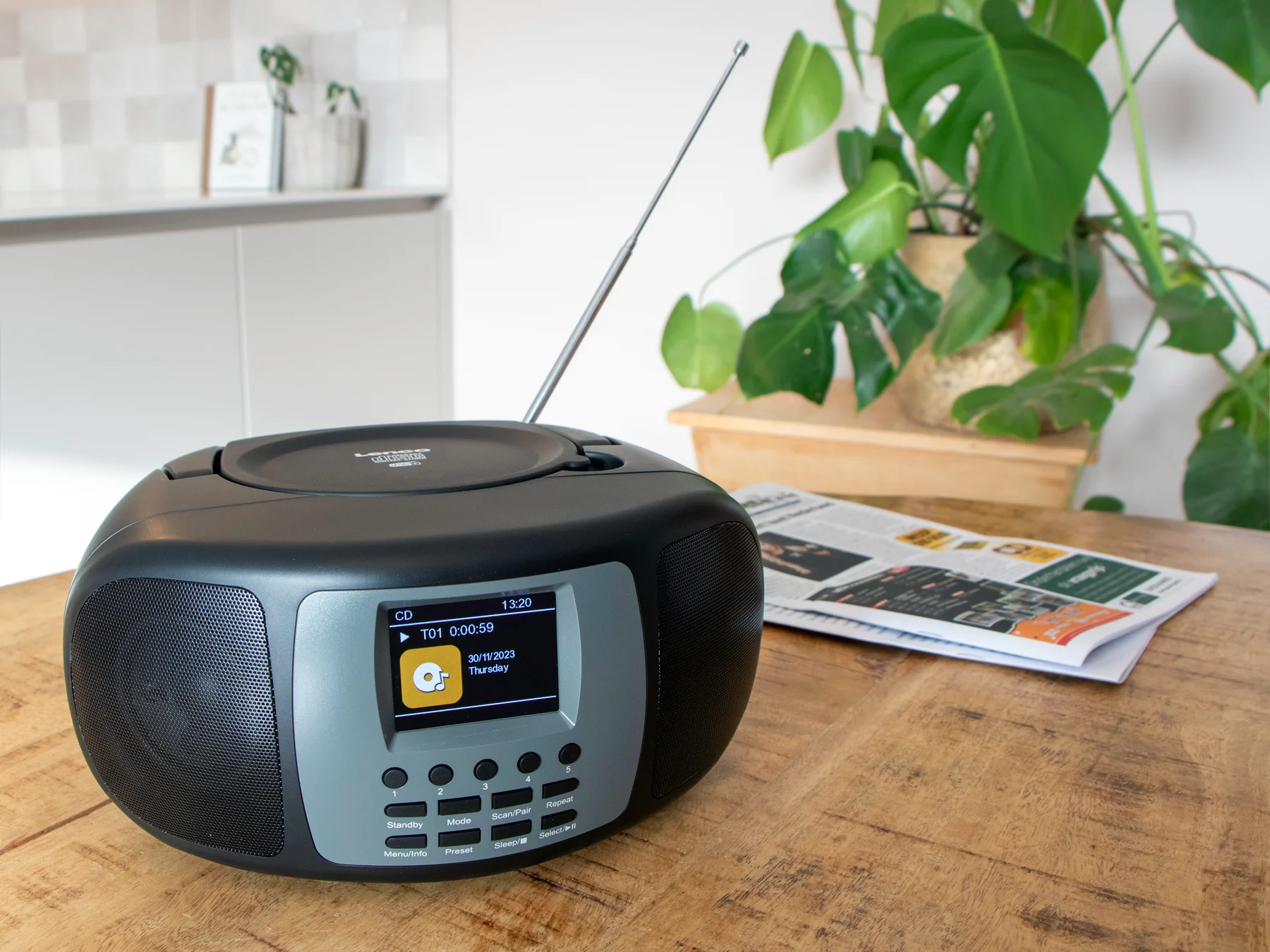 Radio noire avec affichage numérique sur une table en bois. Plante d'intérieur et journal en arrière-plan.