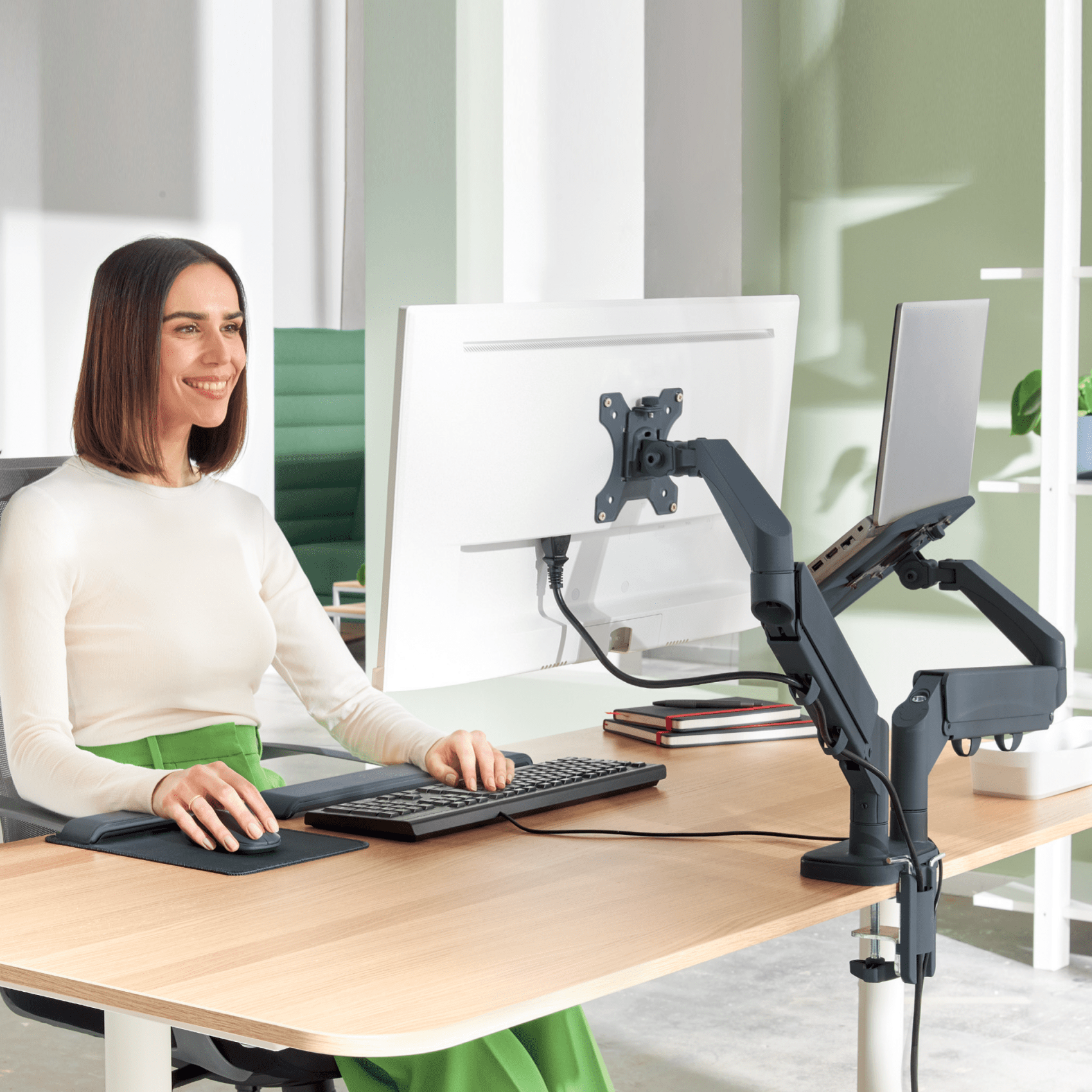 Nő számítógépet használ az íróasztalnál. A monitor és a laptop karra van szerelve.