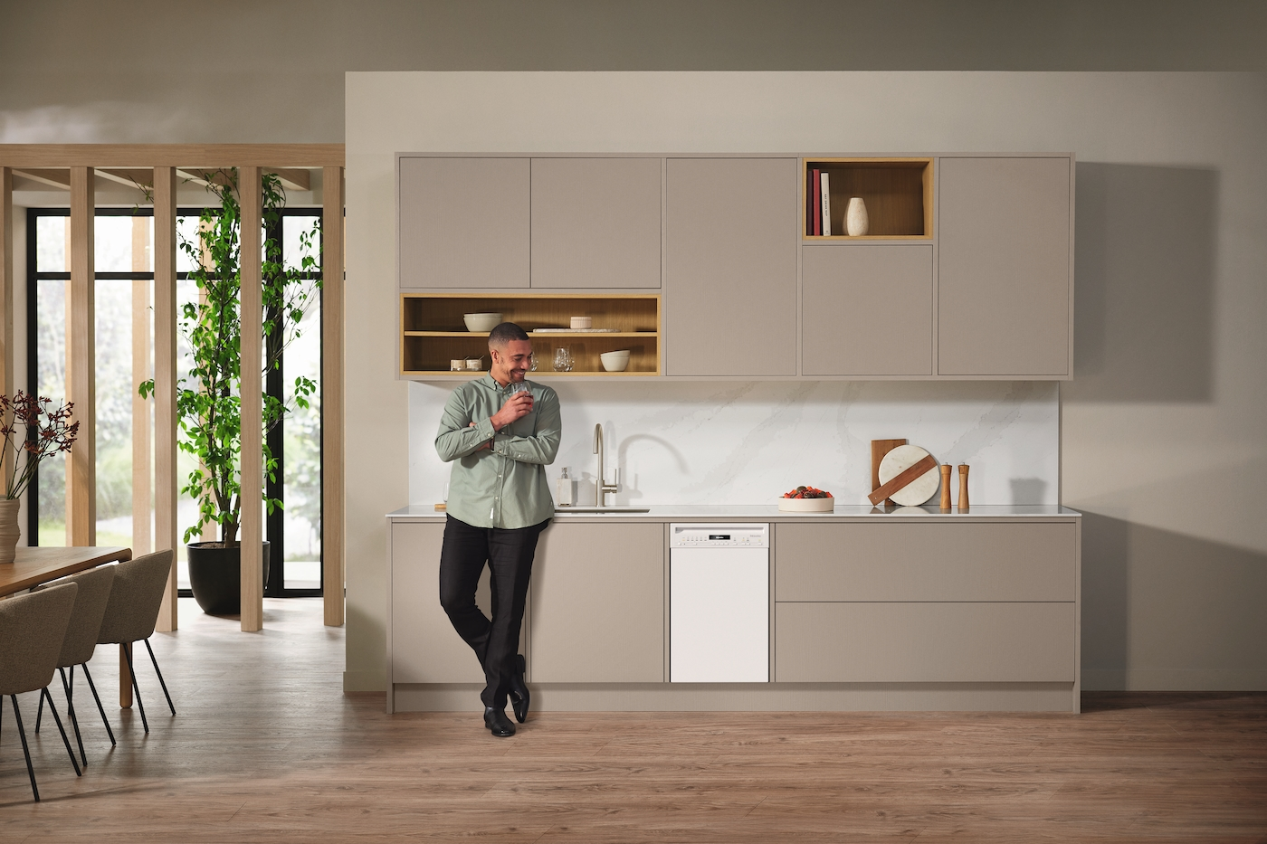 Moderne Küche mit Mann, der sich anlehnt und Glas hält. Weißer Geschirrspüler integriert. Neutrale Schränke und Deko.
