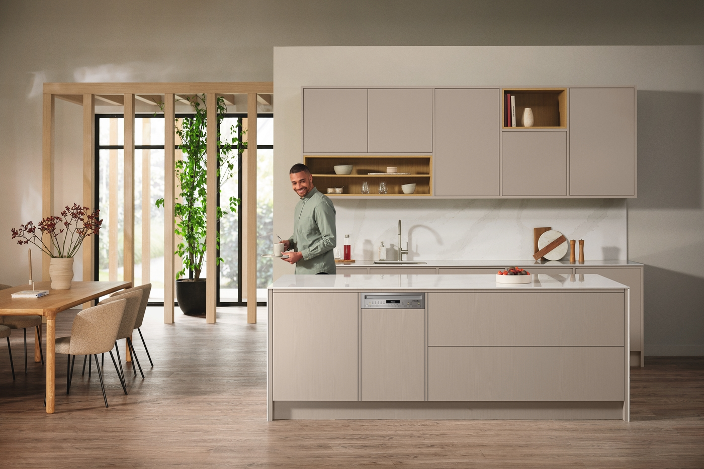 Ein Mann in einer modernen Küche, lächelnd, hält eine Tasse. Die Küche hat beige Schränke und eine große Insel.