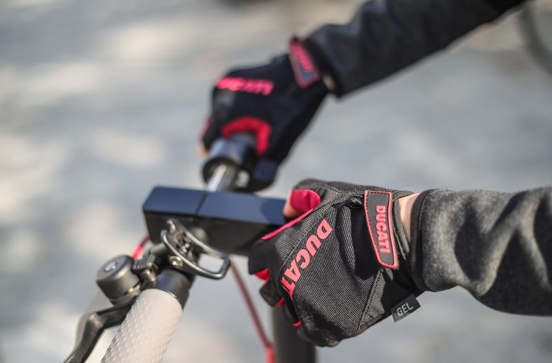 Dłonie w czarnych rękawicach Ducati trzymające kierownicę skutera.