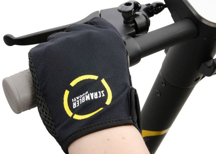 Ręka w rękawicy chwytająca kierownicę skutera. Czarna rękawica ma żółte logo Ducati Scrambler.