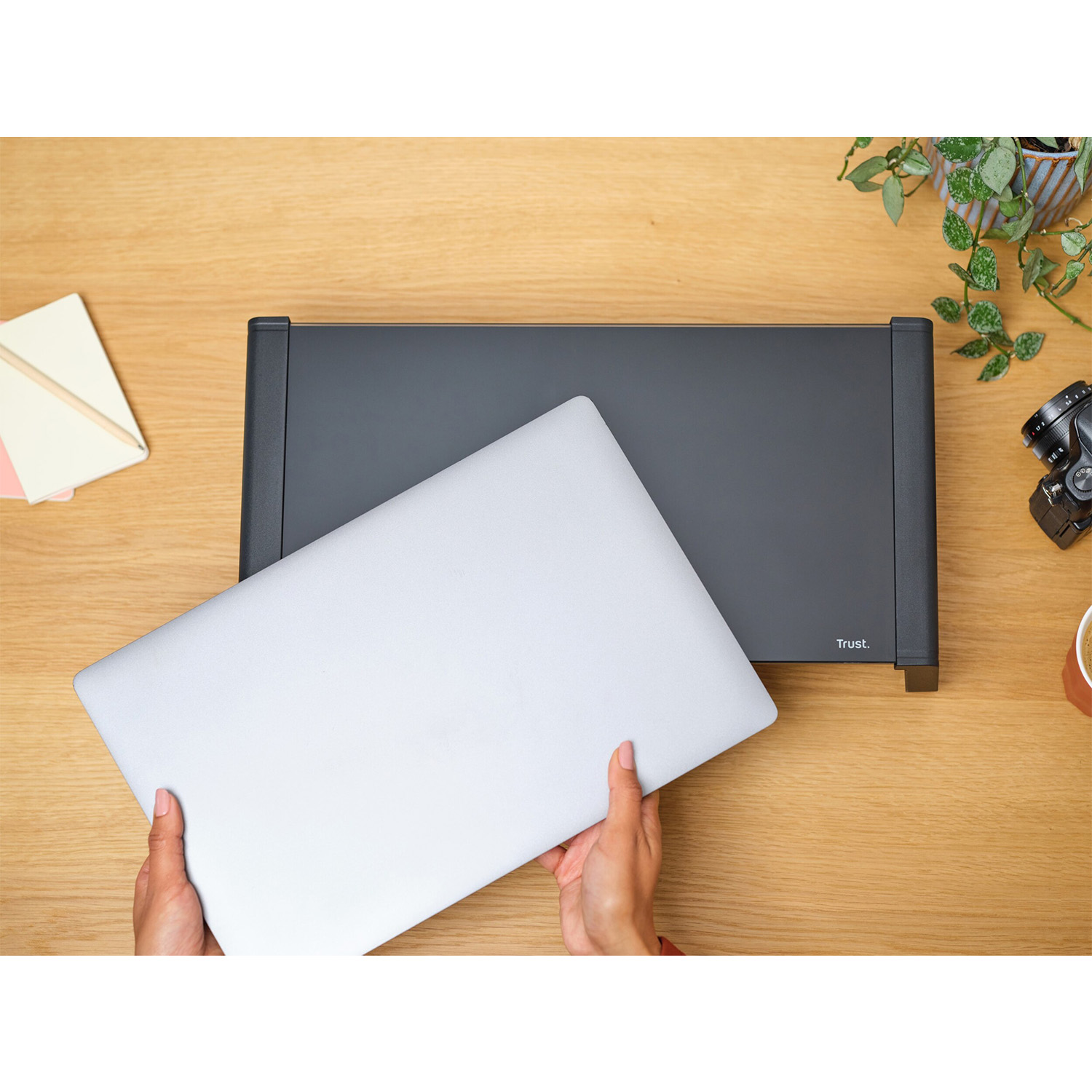 Mani che tengono un laptop su un supporto nero per laptop su una scrivania di legno. Una pianta e una fotocamera sullo sfondo.