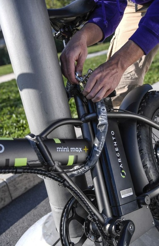 Osoba przypina rower do metalowego słupa. Czarny rower z zielonymi akcentami.