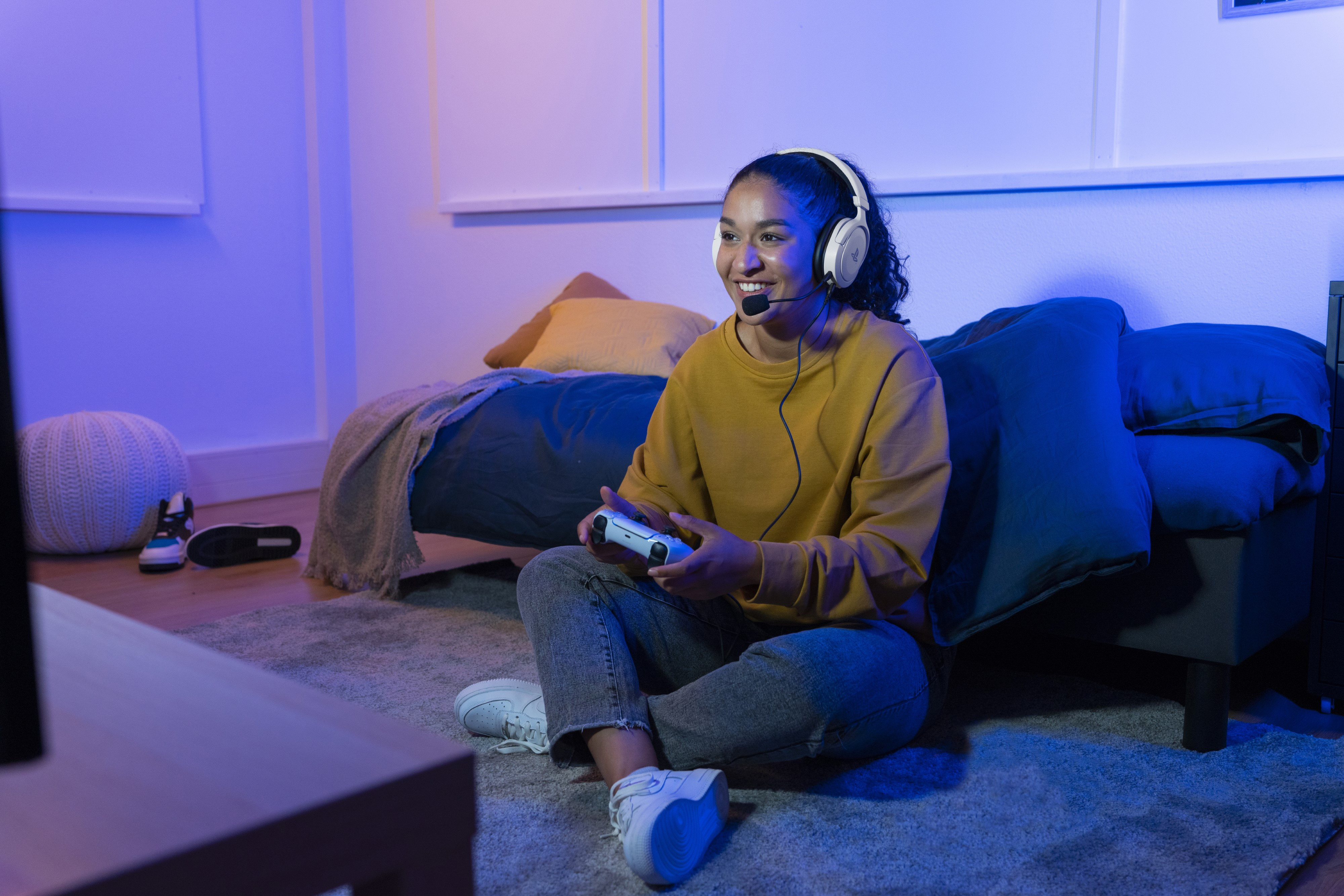 Woman sits gaming with headset and controller. Bedroom has bed, blue/purple lights.