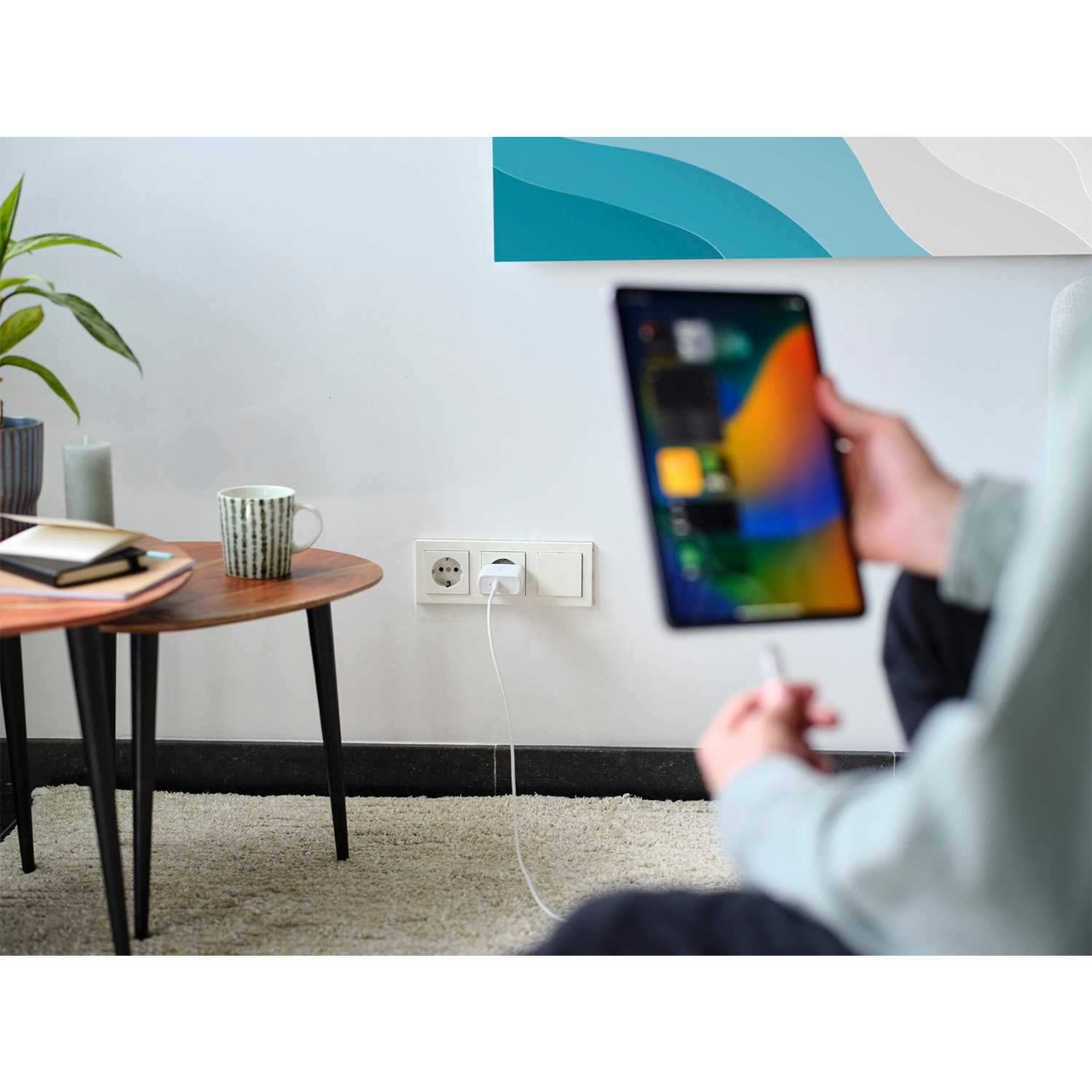 Una persona tiene in mano un tablet. Una spina è collegata al muro. Tazza e libri vicini.