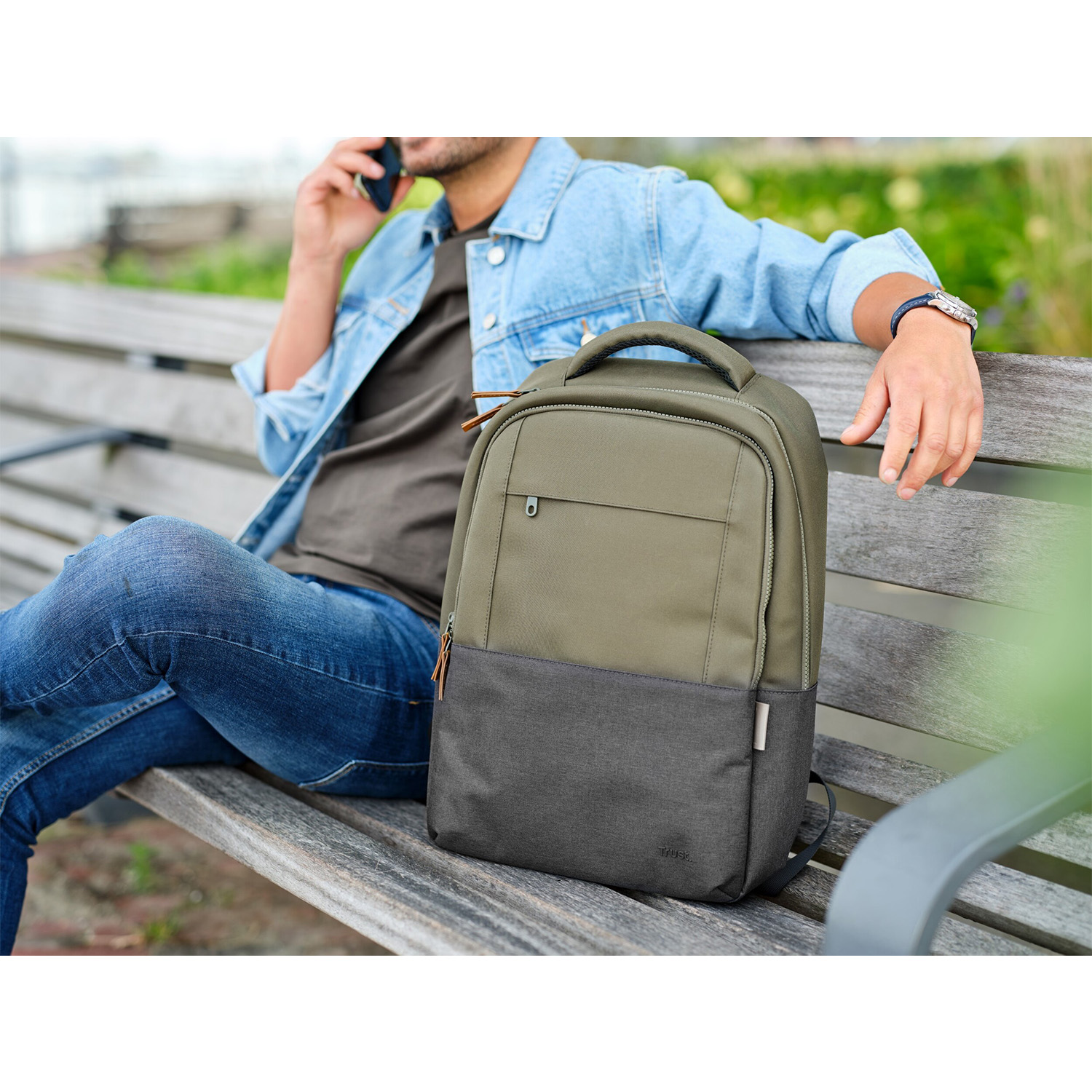 Uomo con zaino su panchina, parla al telefono. Zaino oliva/grigio, jeans, giacca di jeans.