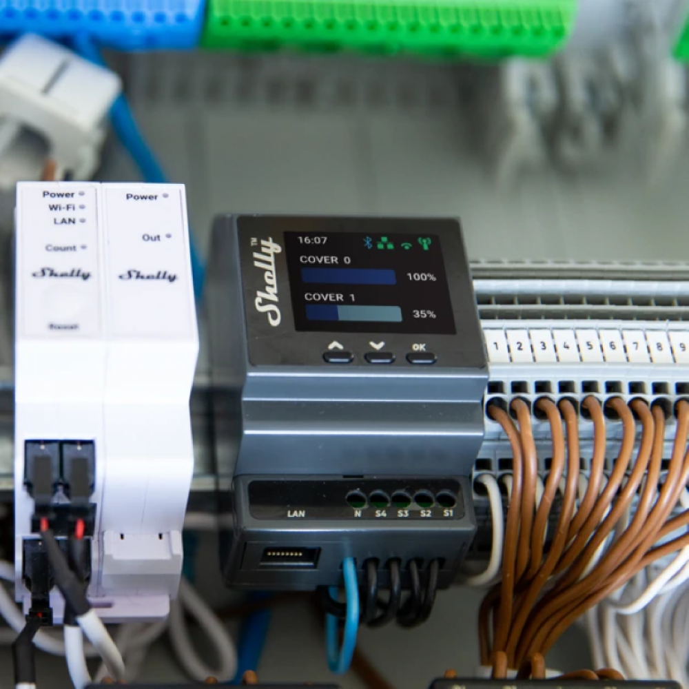 Egy panel egy fehér Shelly eszközzel, egy szürke Shellyvel és elektromos vezetékekkel.