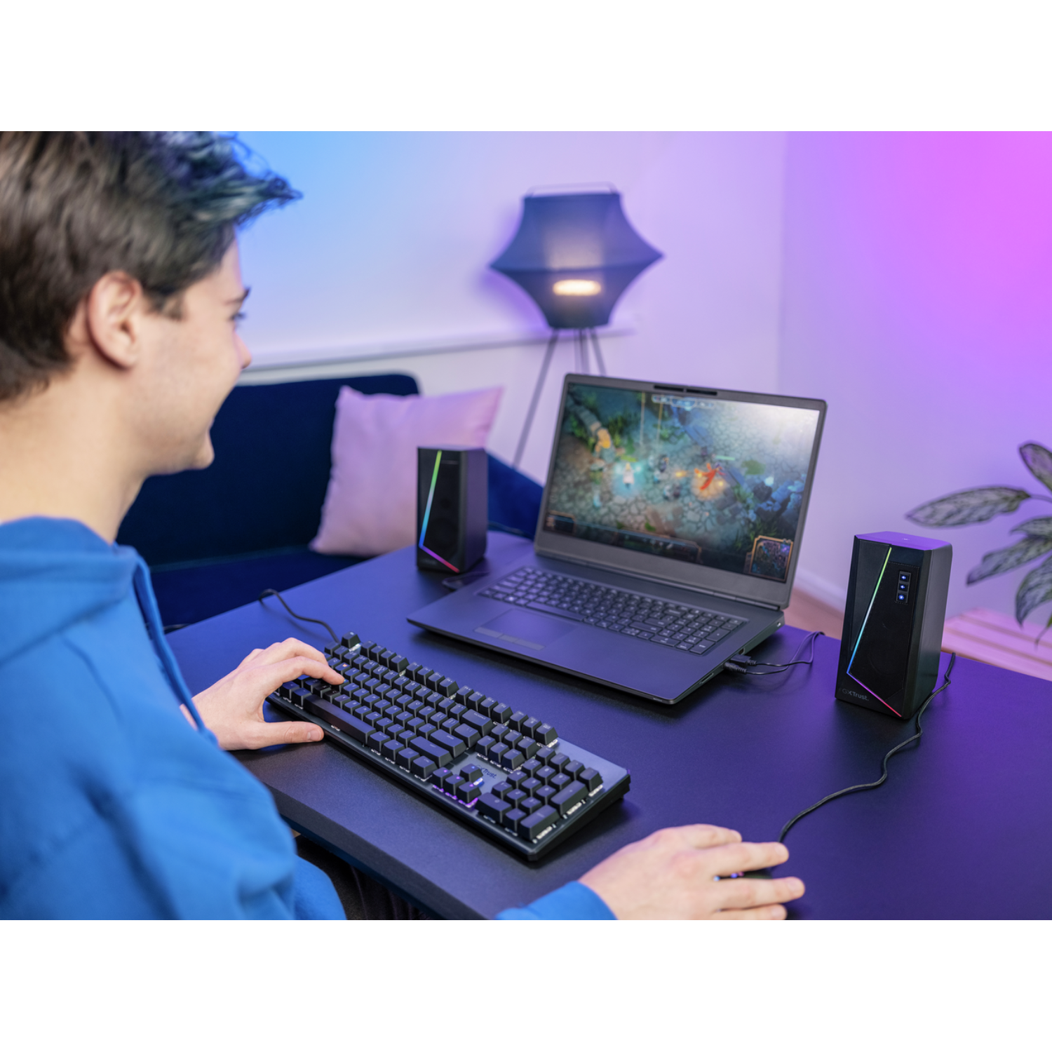 A person gaming with a laptop, keyboard, and speakers on a desk.