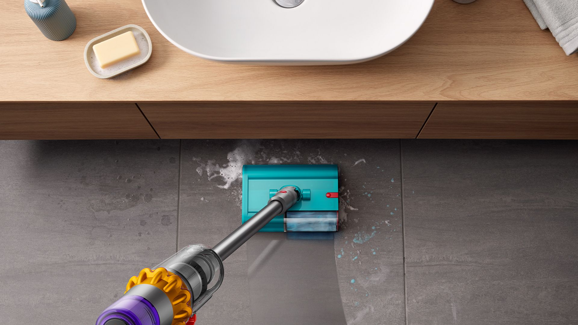 Gri fayans zeminde temizlik yapan turkuaz ve gümüş renkli bir elektrikli süpürge. Arka planda beyaz bir lavabo ve ahşap bir dolap var.