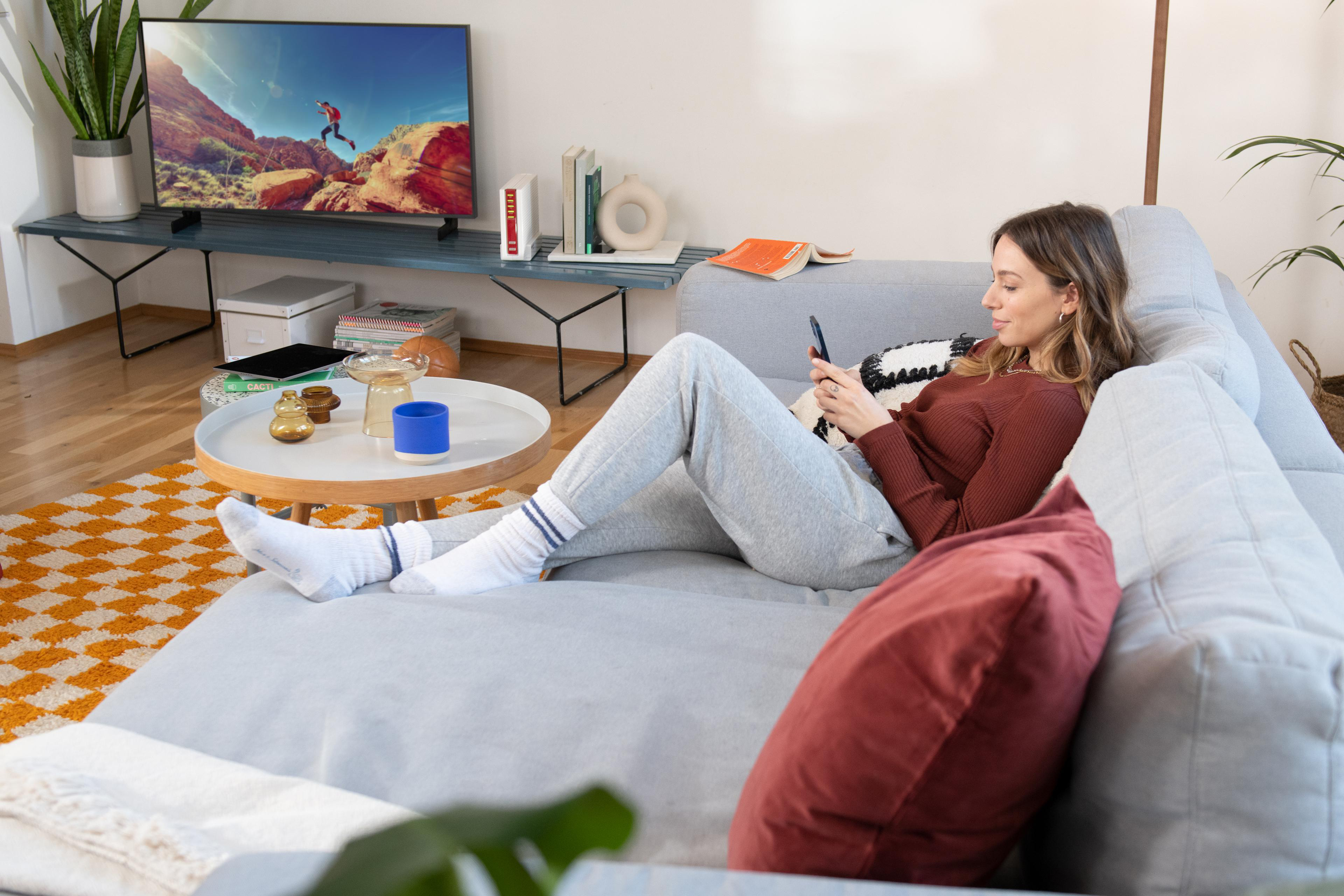 Frau auf grauer Couch, Telefonnutzung, TV schauen, Couchtisch mit Gegenständen und Teppich.